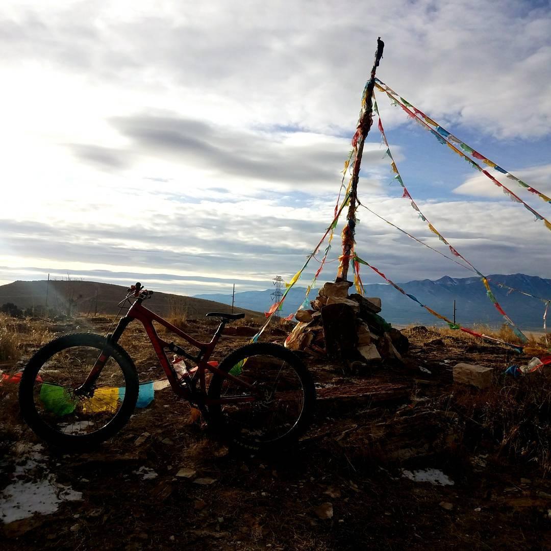 A mountain bike resting on the ground in a rural landscape, with a colorful prayer flag structure and a stone pile in the background. The sky is partly cloudy, revealing a mountainous terrain in the distance. Creed, Roadrunner, Deadwood, Treadstone mountain bike trail.