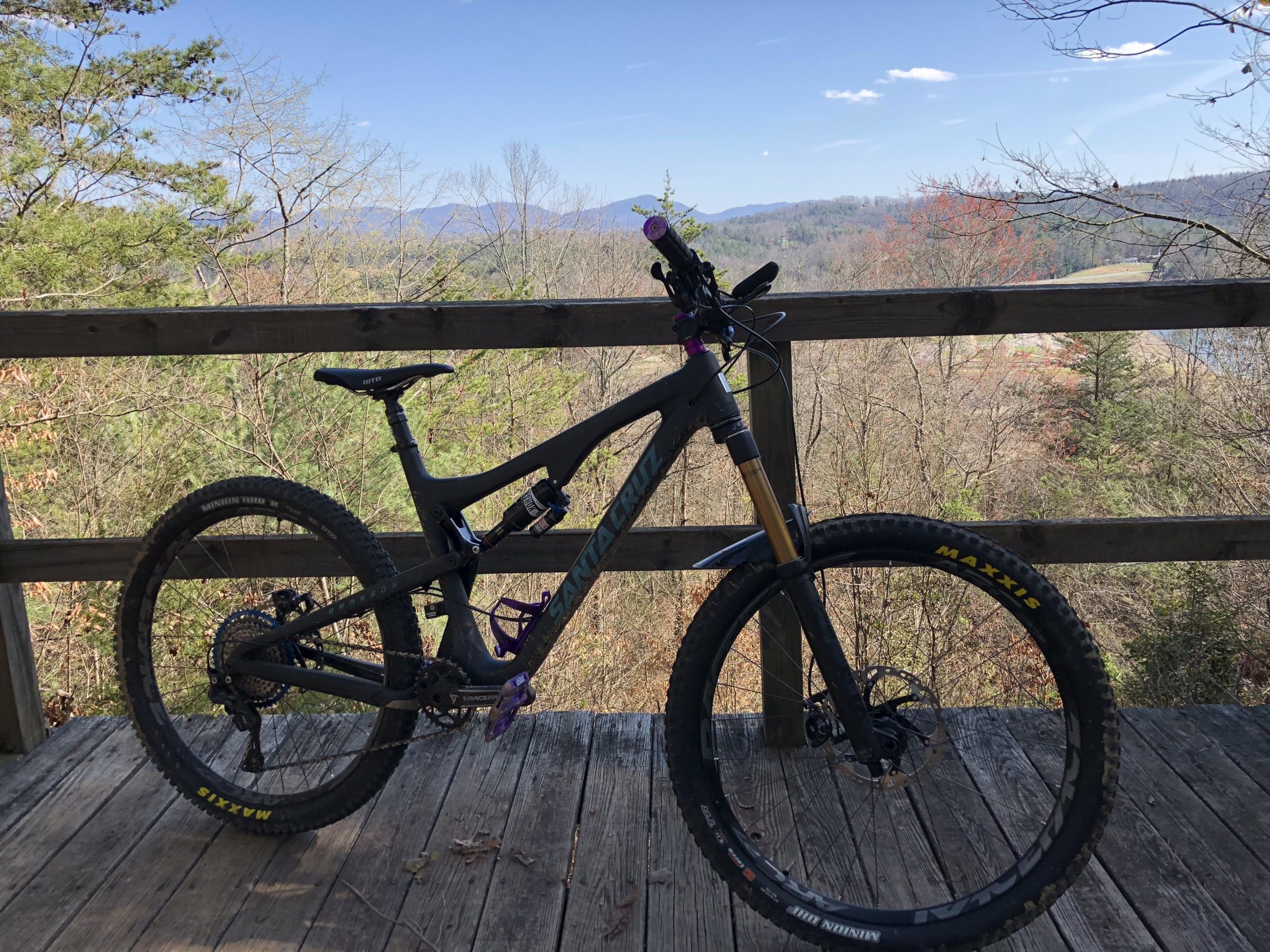 A mountain bike resting on a wooden deck, overlooking a scenic view of trees and rolling hills under a clear blue sky. The bike features a black frame with purple accents and rugged tires. Dark Mountain Trail mountain bike trail.