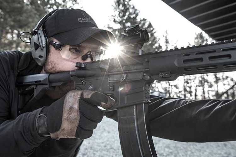 A person in tactical attire aims a rifle at the shooting range, wearing protective ear and eye gear. Sunlight glints off the firearm, and trees are visible in the background.