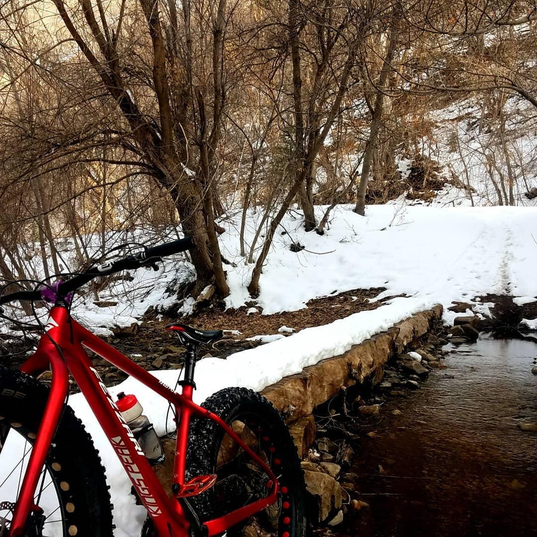 A red mountain bike is parked beside a snow-covered trail leading through a winter landscape, with bare trees in the background. A small creek flows next to a rocky path, partially covered by snow. The scene captures the serene beauty of nature in a cold climate. Whiting Campgrounds mountain bike trail.