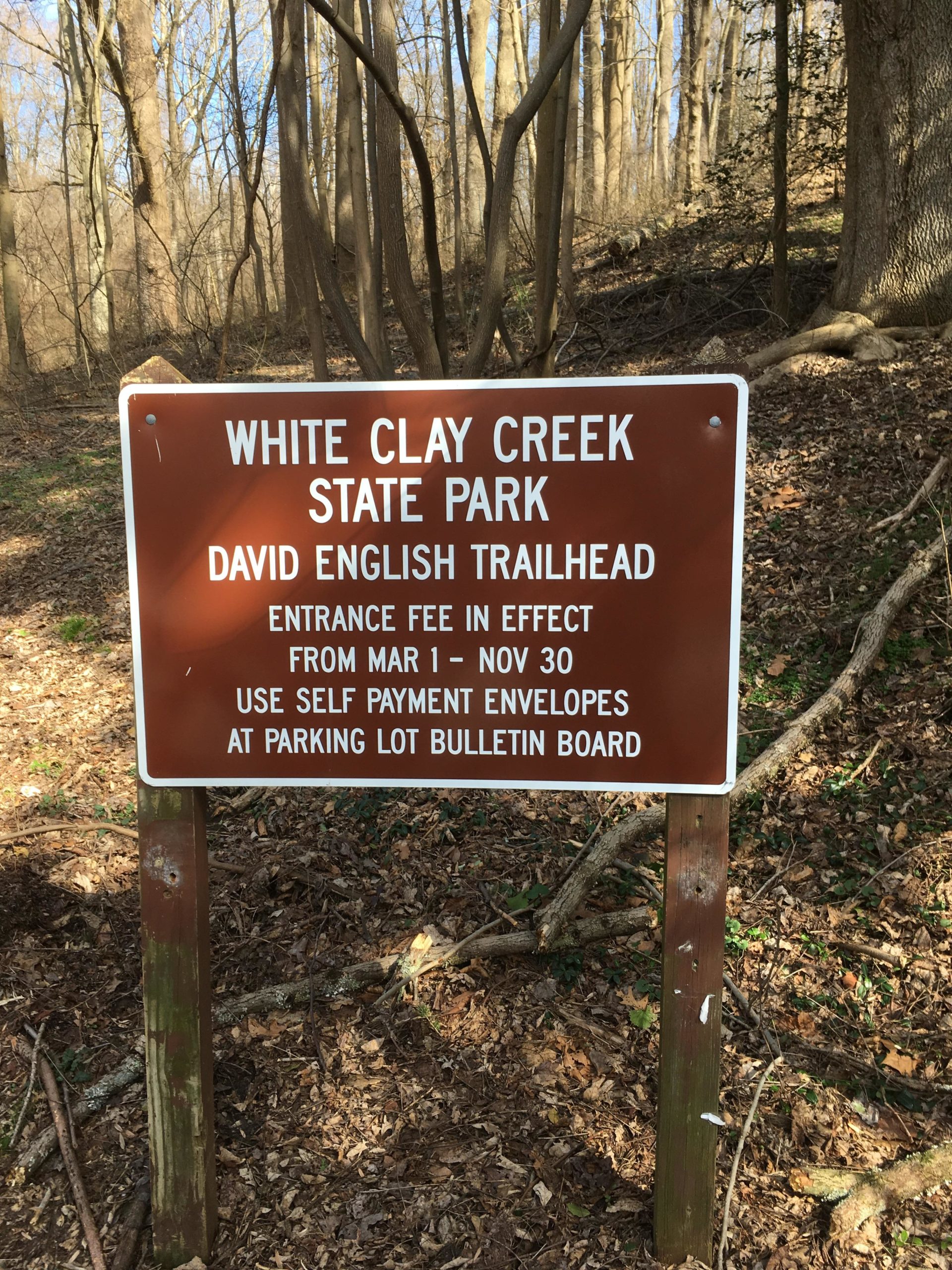 A wooden sign at the David English Trailhead in White Clay Creek State Park, displaying information about the entrance fee in effect from March 1 to November 30 and instructions for self-payment using envelopes available at the parking lot bulletin board. The background features a wooded area with trees and fallen leaves. White Clay Creek mountain bike trail.