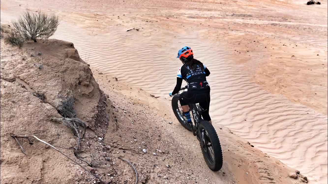 A person riding a fat bike down a sandy trail, with rippled sand patterns in the background and sparse vegetation on the rocky terrain. The rider is wearing a helmet and cycling gear. Mariposa Fat Bike Trails mountain bike trail.