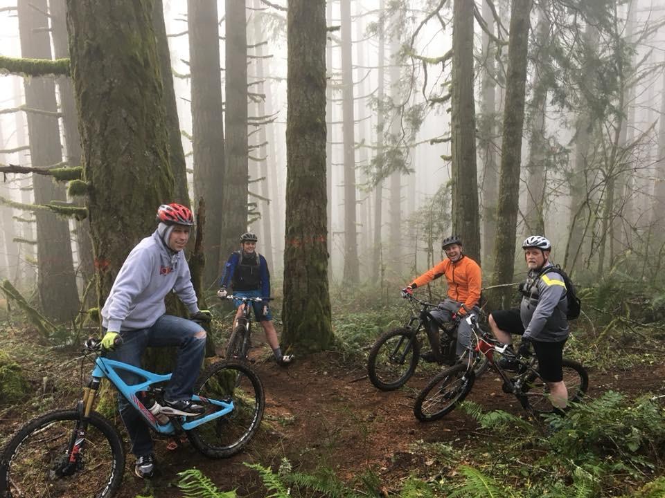 Four mountain bikers are gathered in a misty forest, surrounded by tall trees. Two of the riders are seated on their bikes, while the other two stand nearby. The first rider is wearing a gray sweatshirt and a red helmet, the second is in an orange jacket, and the third is in a gray shirt with black shorts. The atmosphere is foggy, adding a sense of adventure to their outdoor setting. Ferns and underbrush can be seen on the forest floor. Whypass mountain bike trail.