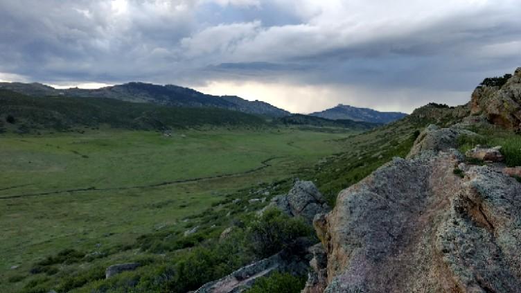 A panoramic view of a valley surrounded by rolling hills and rocky formations under a cloudy sky. The landscape features lush green grass and patches of shrubs, creating a serene natural setting. Coyote Ridge mountain bike trail.