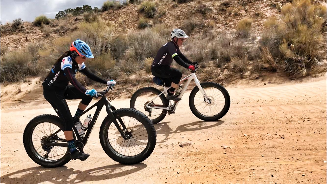 Two cyclists ride along a sandy, off-road trail surrounded by sparse vegetation and hills. One cyclist, wearing a blue helmet and a black jersey with blue accents, is on a black bicycle with thick tires. The other cyclist, dressed in a dark jersey and pants, rides a white bicycle with a slightly different style. The scene captures the action and enjoyment of mountain biking in a natural outdoor setting. Mariposa Fat Bike Trails mountain bike trail.