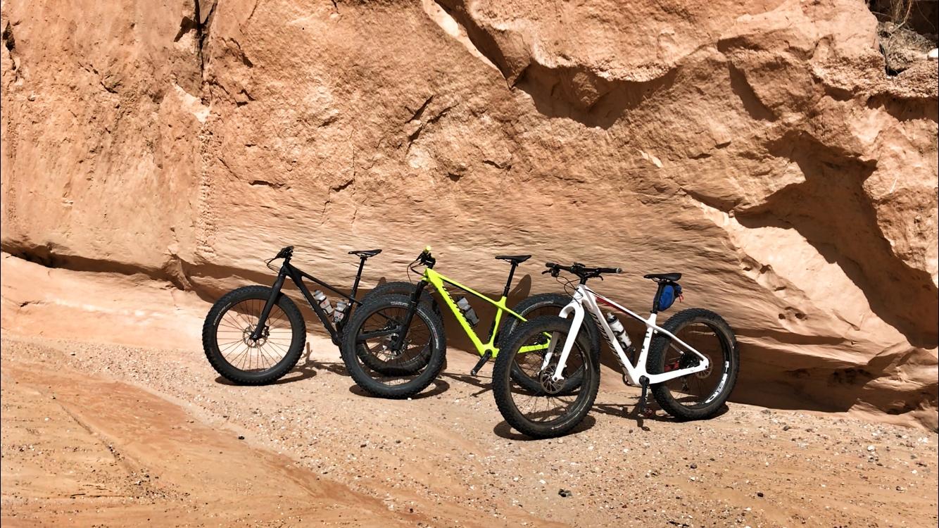 Three fat bikes of different colors—black, bright green, and white—are parked against a textured, sandy rock face in a desert setting. The ground is dirt, and the scene suggests a sunny outdoor location. Mariposa Fat Bike Trails mountain bike trail.