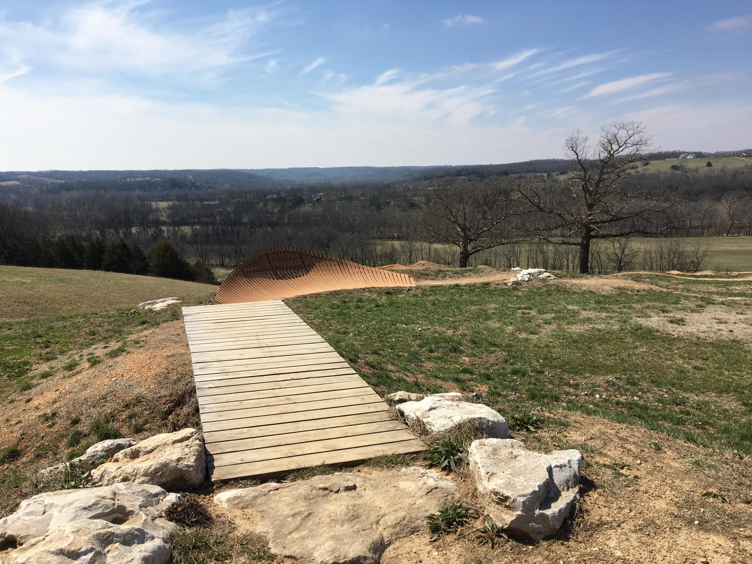 A wooden pathway leads through a grassy landscape, with a large, curved sculpture made of metal towards the horizon. The scene features rolling hills in the background under a bright blue sky with wispy clouds. Leafless trees are scattered across the fields, adding to the serene, open countryside ambiance. Two Rivers Bike Park mountain bike trail.