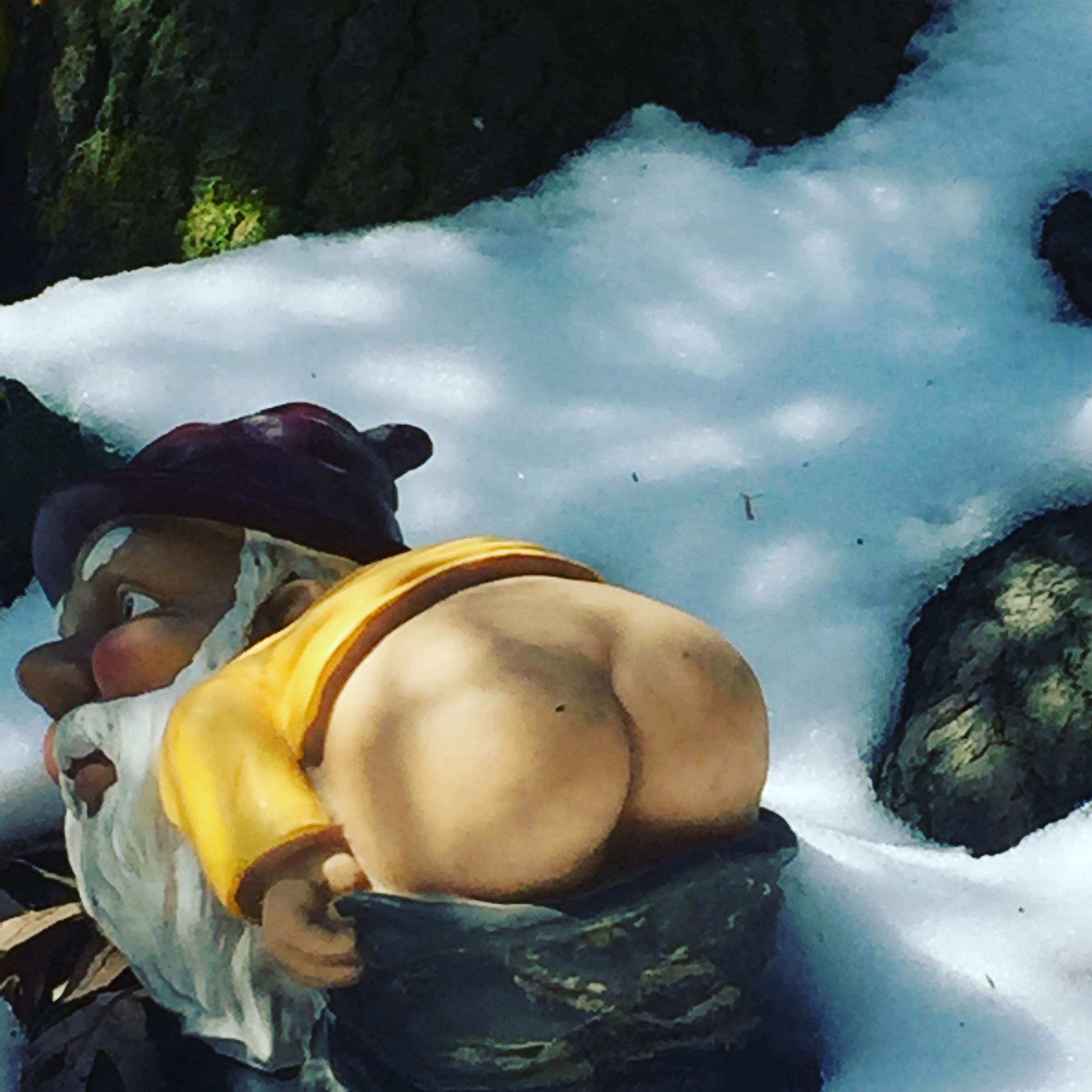 A humorous garden gnome figurine bending over, partially buried in snow, with a bright yellow shirt and a purple hat, surrounded by tree roots and snow. Mohican mountain bike trail.