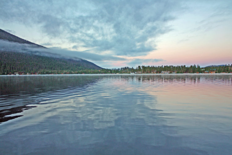 A tranquil lake scene at dusk, featuring calm waters reflecting the soft colors of the sky with clouds and a wooded mountain in the background. The shoreline reveals a mix of trees and distant houses.