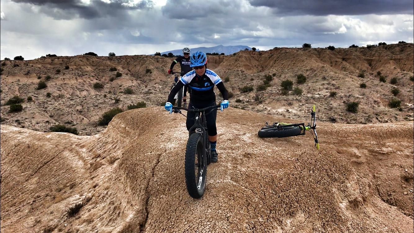 A mountain biker navigating a rocky terrain, wearing a blue and black jersey and helmet. In the background, another cyclist is visible on a similar track. The landscape features dry, uneven ground with sparse vegetation under a cloudy sky. A second bicycle lies on its side nearby. Mariposa Fat Bike Trails mountain bike trail.