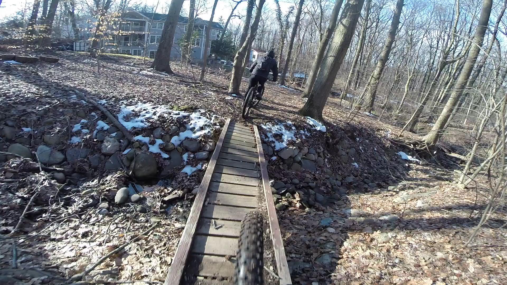 A mountain biker navigating a narrow wooden bridge over a rocky area in a forested environment, with scattered leaves and patches of snow on the ground. In the background, a residential building is visible among the trees. Richmond Avenue and Forest Hill road mountain bike trail.
