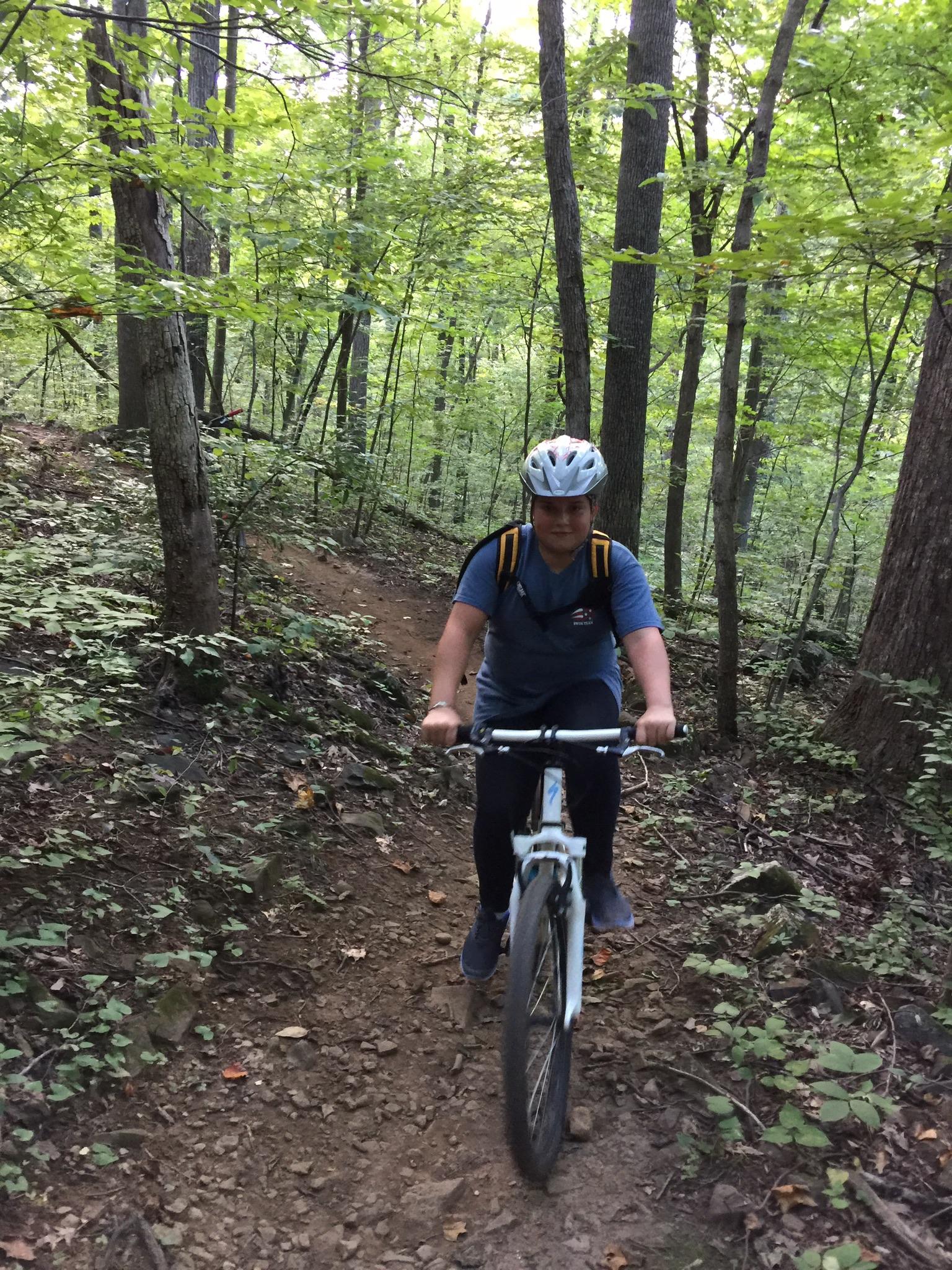A person riding a mountain bike on a narrow, rocky trail surrounded by lush green trees in a forested area. The rider is wearing a helmet and a backpack, indicating safety and preparedness for outdoor biking. Conway Robinson State Forest mountain bike trail.
