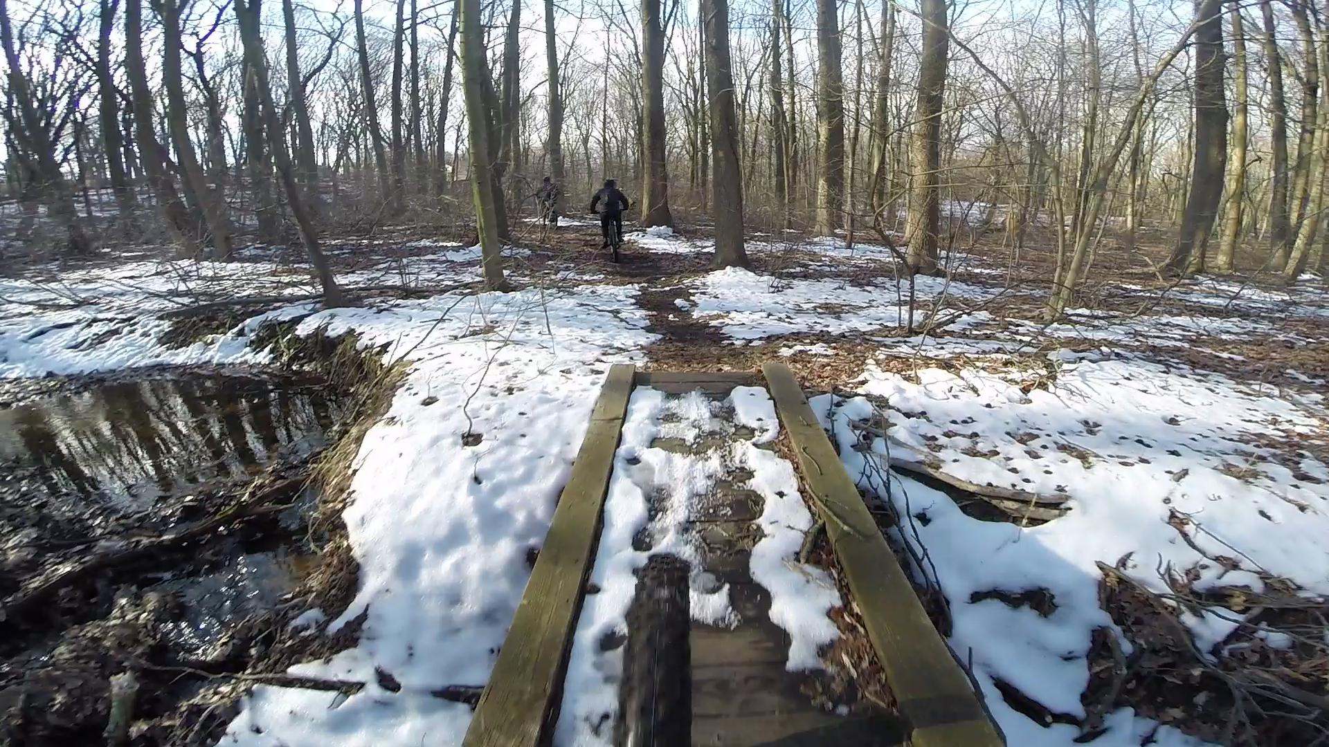 A path through a forest covered with patches of snow and fallen leaves, featuring a wooden bridge over a small stream. In the background, a person is riding a bicycle along the trail amidst the trees. The scene is set in a winter environment, showcasing bare branches and bright sunlight filtering through. Richmond Avenue and Forest Hill road mountain bike trail.