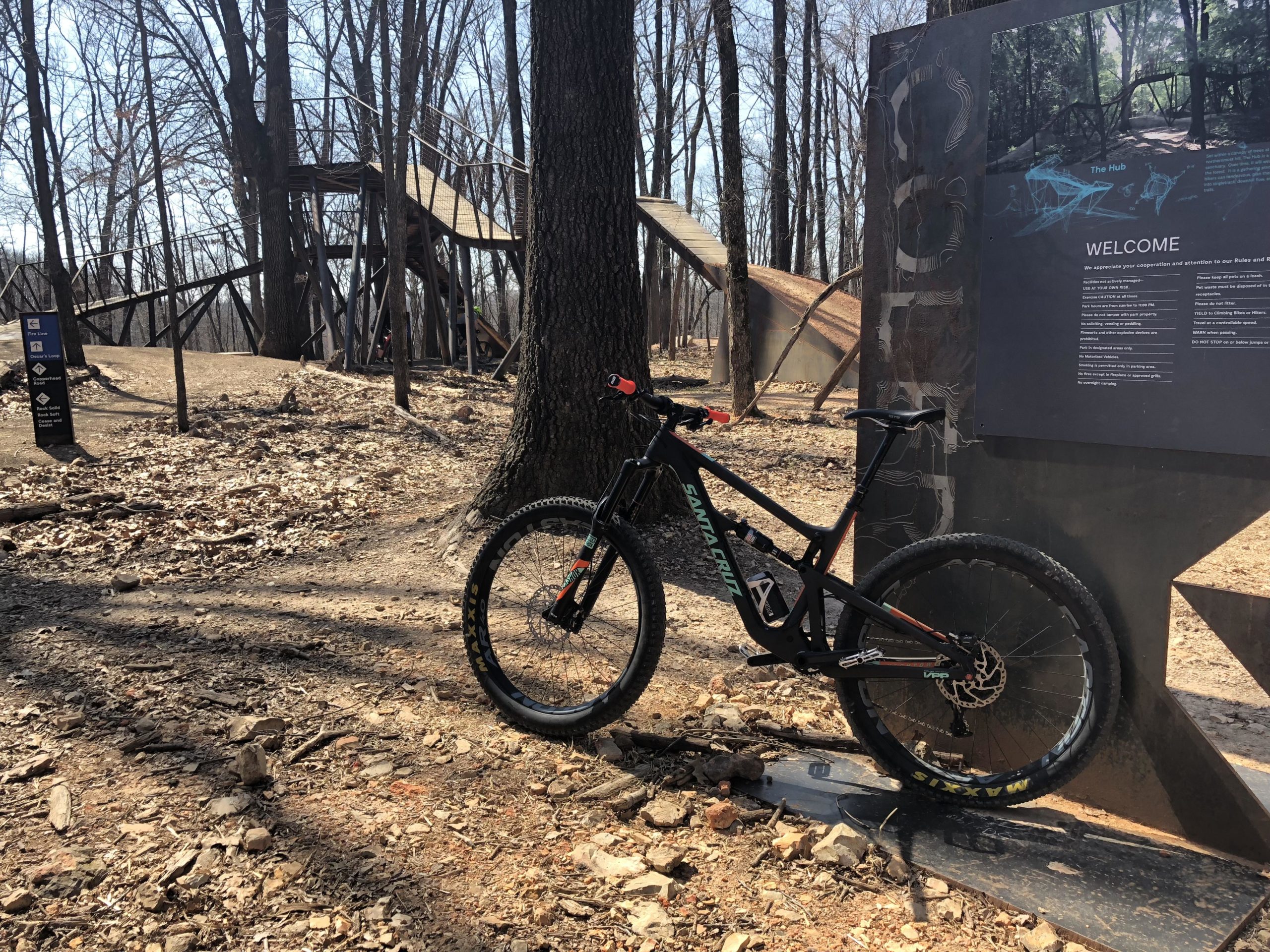 Mountain bike leaning against an information sign in a wooded area, featuring a dirt trail and a wooden structure in the background. The scene is set in a natural environment with bare trees and leaf litter on the ground. Coler Mountain Bike Preserve mountain bike trail.