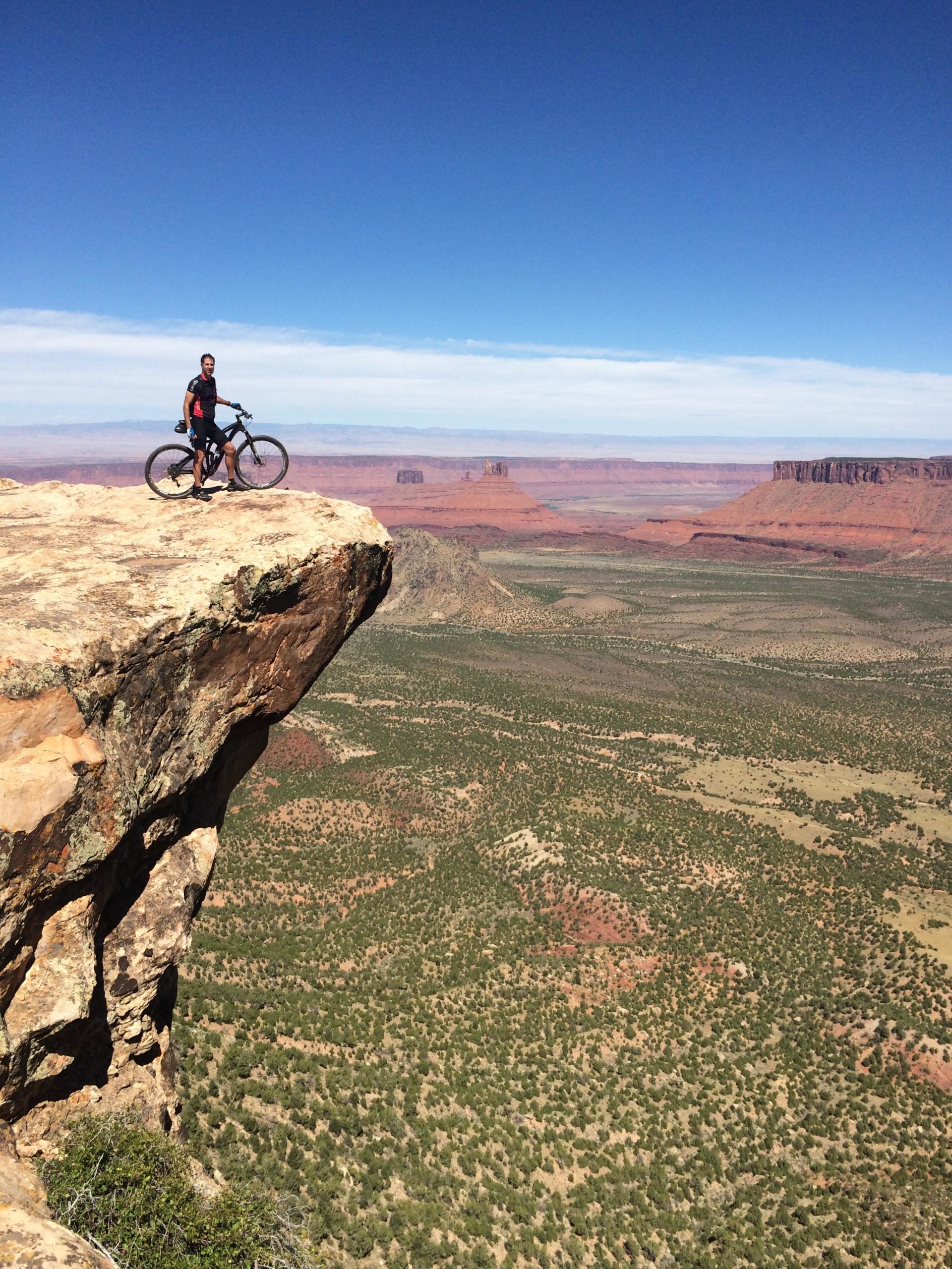 A mountain biker stands on the edge of a rocky cliff, overlooking a vast desert landscape filled with greenery and red rock formations under a clear blue sky. The Whole Enchilada mountain bike trail.