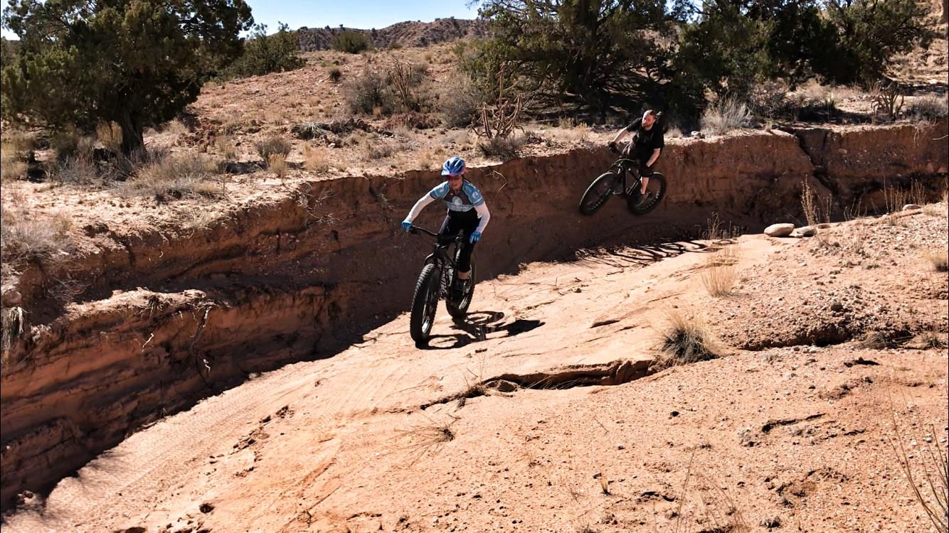Two cyclists maneuvering through a dry, rocky landscape. One rider, dressed in a blue and white jersey, is navigating a steep, sandy slope, while the other rider, wearing a black outfit, is performing a jump off a ledge. The scene is set in a rugged outdoor area with sparse vegetation and clear blue skies. Mariposa Fat Bike Trails mountain bike trail.