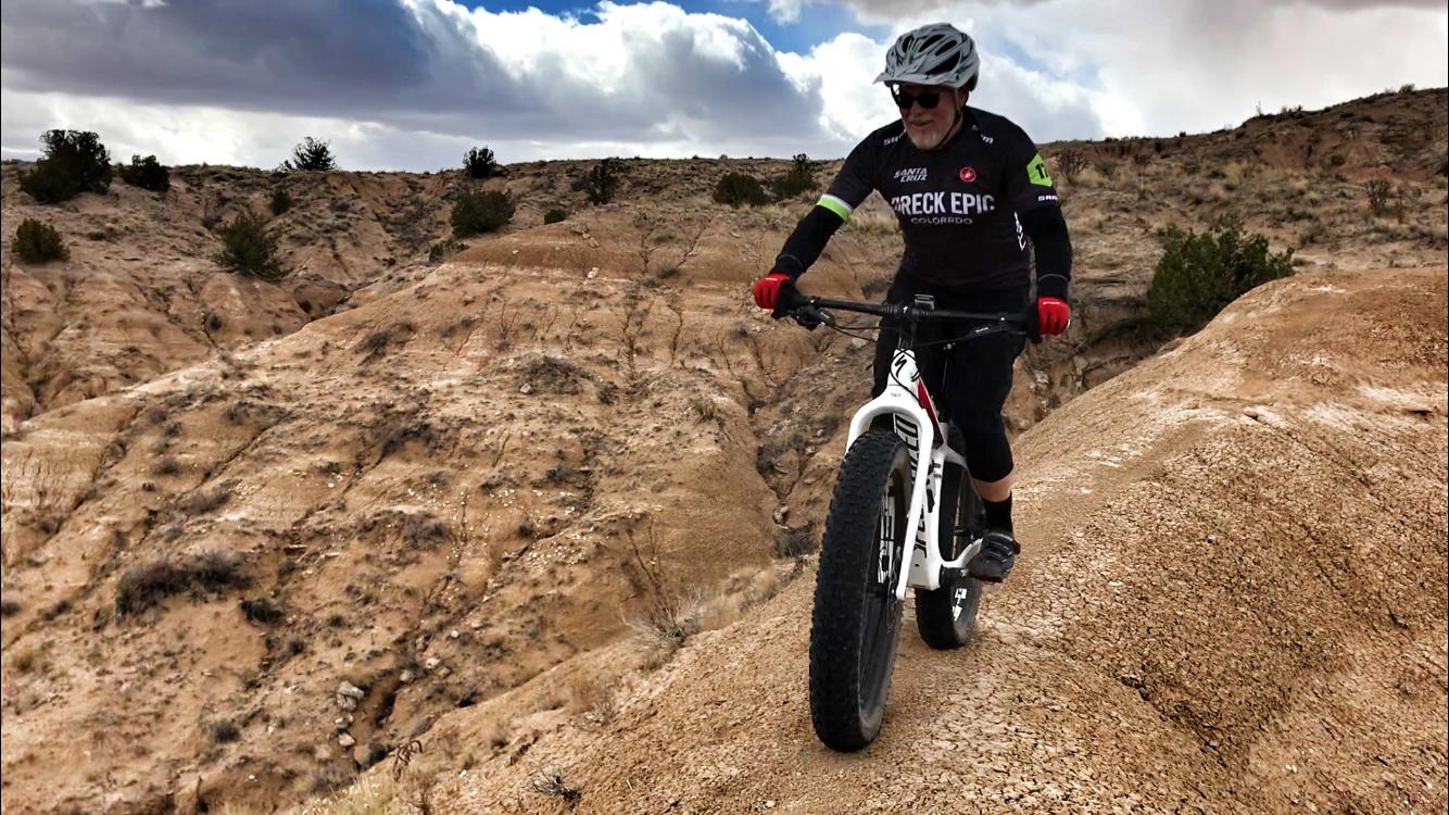 A person riding a fat bike on a rugged dirt trail with hilly terrain, wearing a helmet and cycling gear. The landscape features dry, rocky surroundings under a partly cloudy sky. Mariposa Fat Bike Trails mountain bike trail.