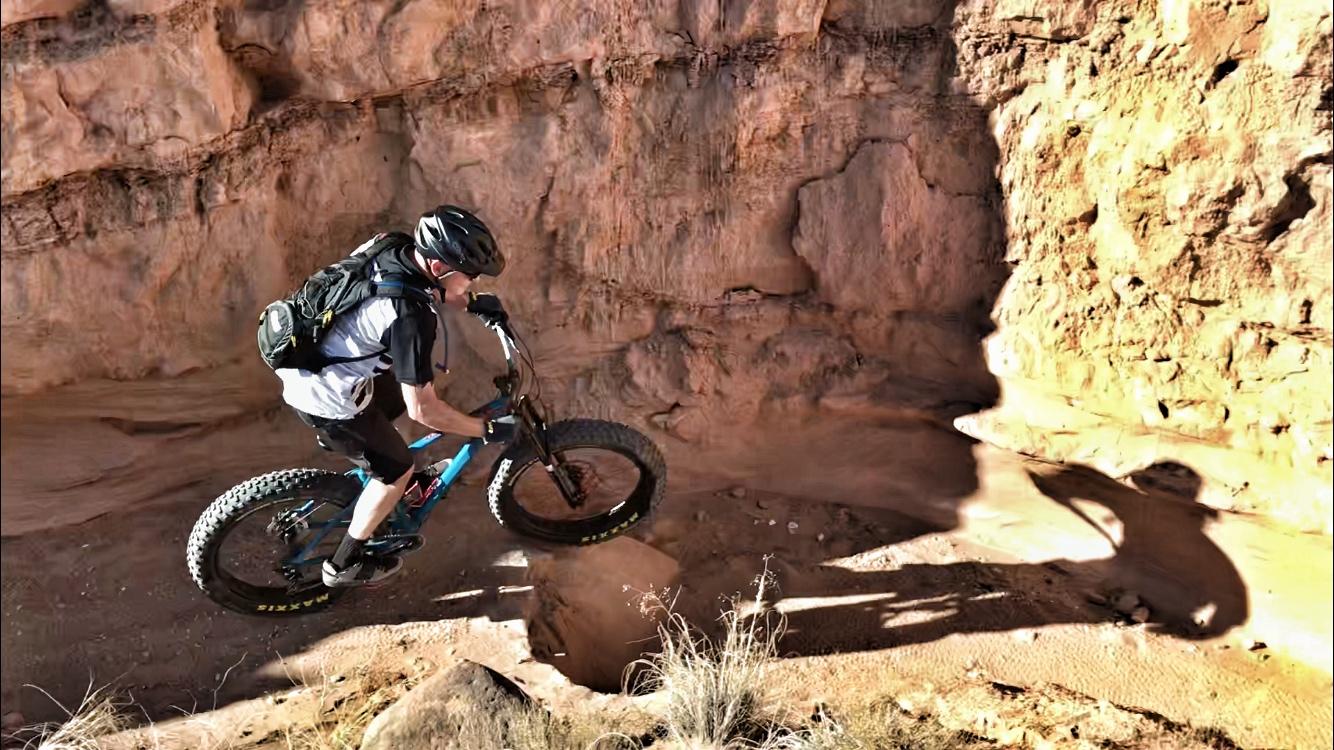A mountain biker jumping over a rocky terrain in a canyon. The rider is wearing a helmet and a backpack, captured mid-air with a dynamic pose against a backdrop of rugged, earthy rock formations. Mariposa Fat Bike Trails mountain bike trail.