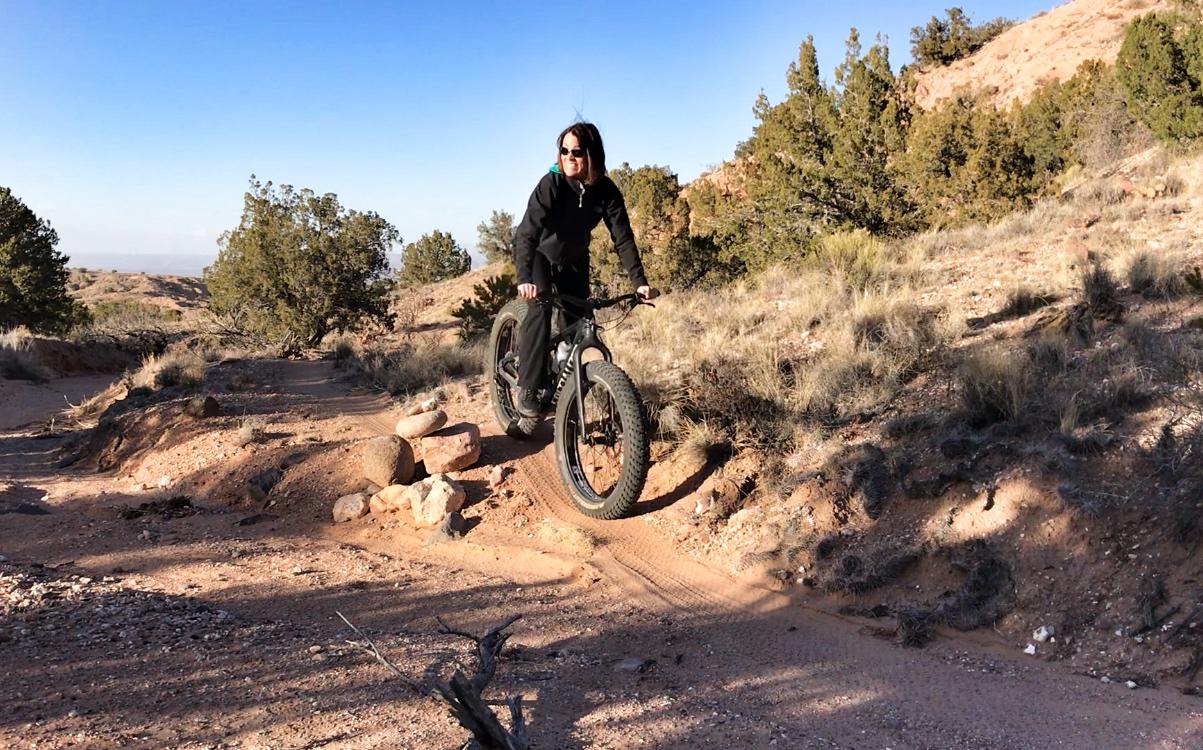 A person riding a fat bike on a rocky trail in a natural outdoor setting, surrounded by scrub vegetation and hills under a clear blue sky. Mariposa Fat Bike Trails mountain bike trail.