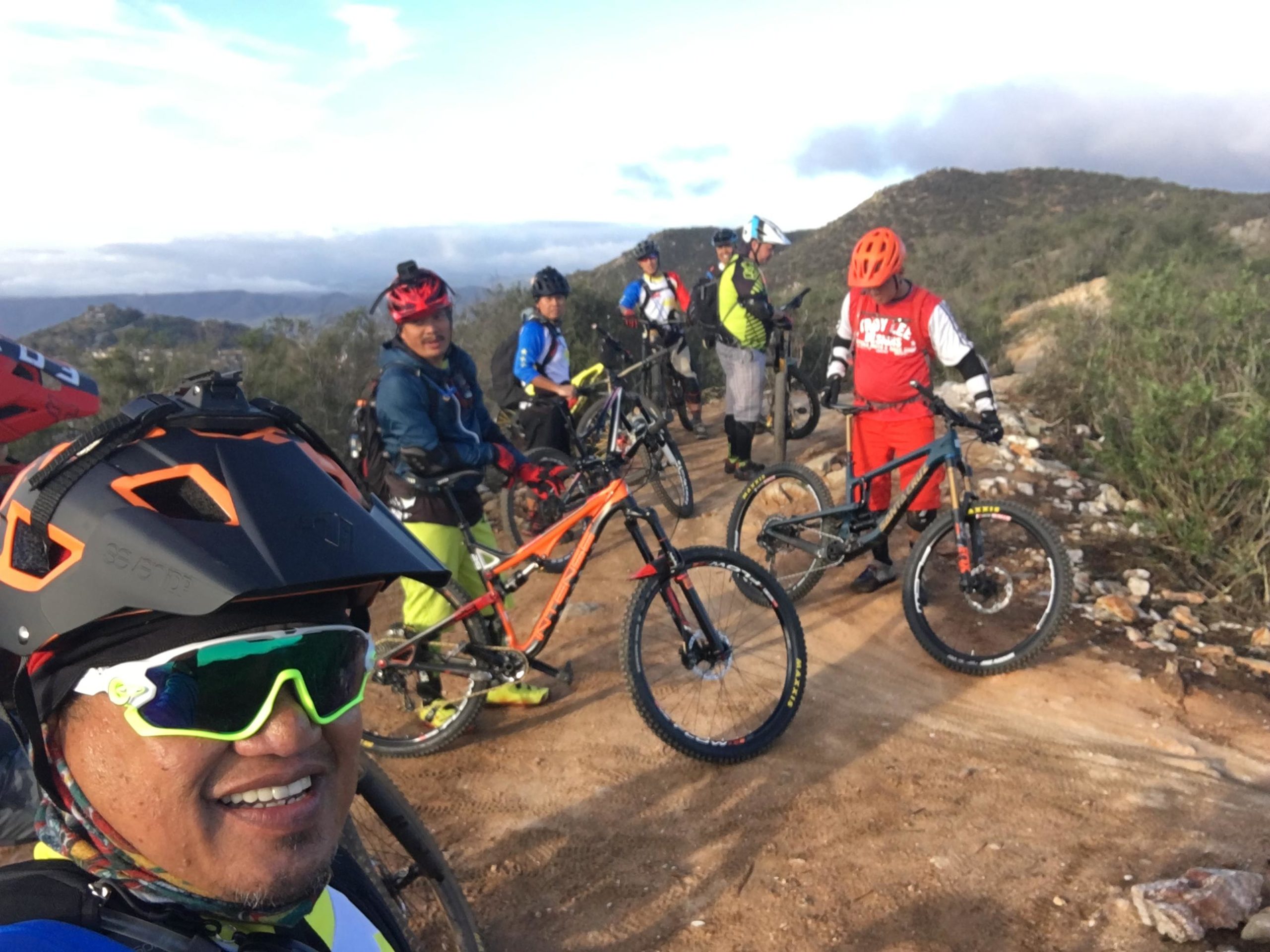 A group of six mountain bikers poses on a dirt trail surrounded by hills and vegetation. The riders are wearing colorful biking gear and helmets, with various mountain bikes beside them. The scene captures a sunny day with blue skies and clouds in the background. One rider is taking a selfie while smiling at the camera. The Nursery mountain bike trail.