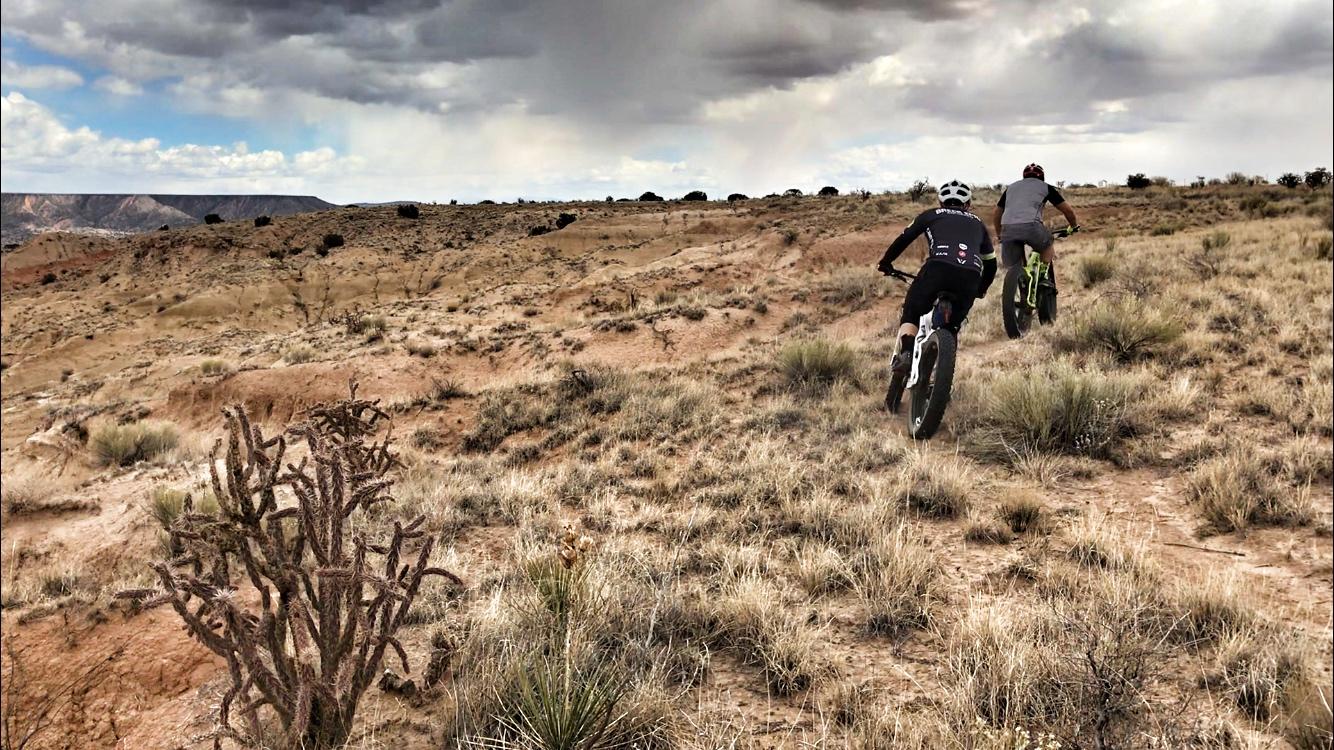 Two mountain bikers ride along a rugged trail in a desert landscape, surrounded by dry grass and sparse vegetation. A cactus is visible in the foreground, and dark clouds fill the sky, suggesting an impending storm. The terrain features hills and distant rocky formations. Mariposa Fat Bike Trails mountain bike trail.