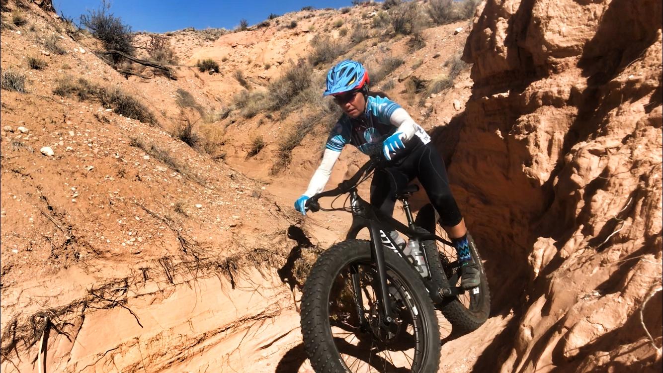A cyclist navigating a rocky trail on a fat bike in a desert landscape, surrounded by red dirt and sparse vegetation. The cyclist is wearing a blue helmet and gloves, focused on maintaining balance while riding along a narrow path. Mariposa Fat Bike Trails mountain bike trail.