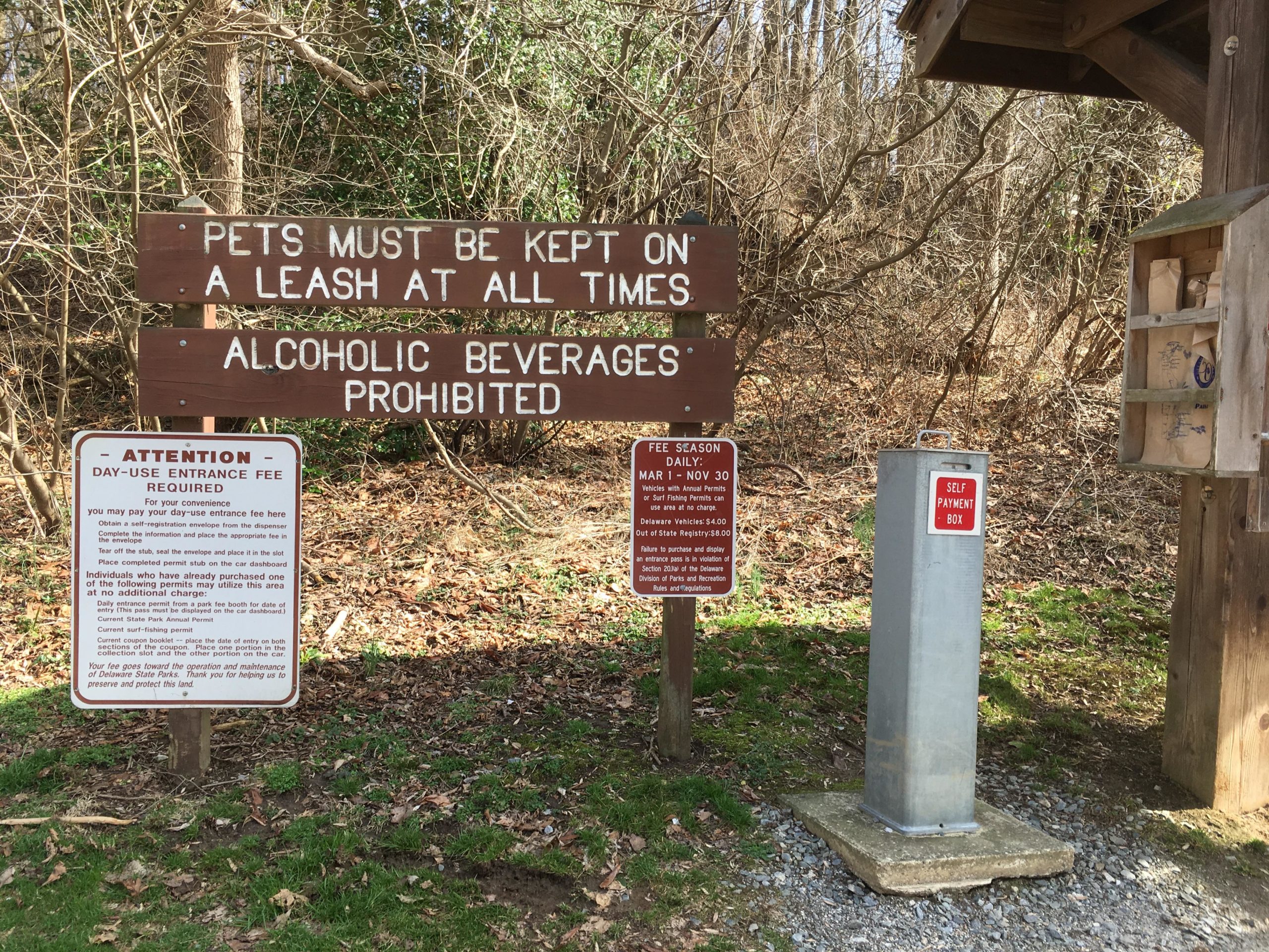 A wooden sign indicating park rules, stating that pets must be kept on a leash at all times and that alcoholic beverages are prohibited. Below, there is a notice about a day-use entrance fee, along with instructions for payment. A metal self-payment box is visible nearby, alongside a small wooden information box containing brochures. The surrounding area features trees and a gravel path. White Clay Creek mountain bike trail.