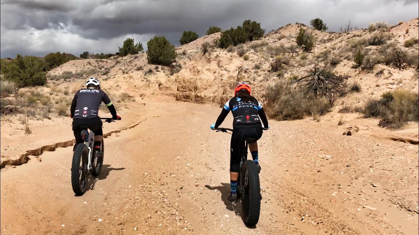Two mountain bikers navigate a sandy trail surrounded by rugged terrain and sparse vegetation under a partly cloudy sky. One rider wears a helmet and a dark jersey, while the other is in a bright orange helmet and a blue accent jersey, both riding fat bikes on a rocky path. Mariposa Fat Bike Trails mountain bike trail.