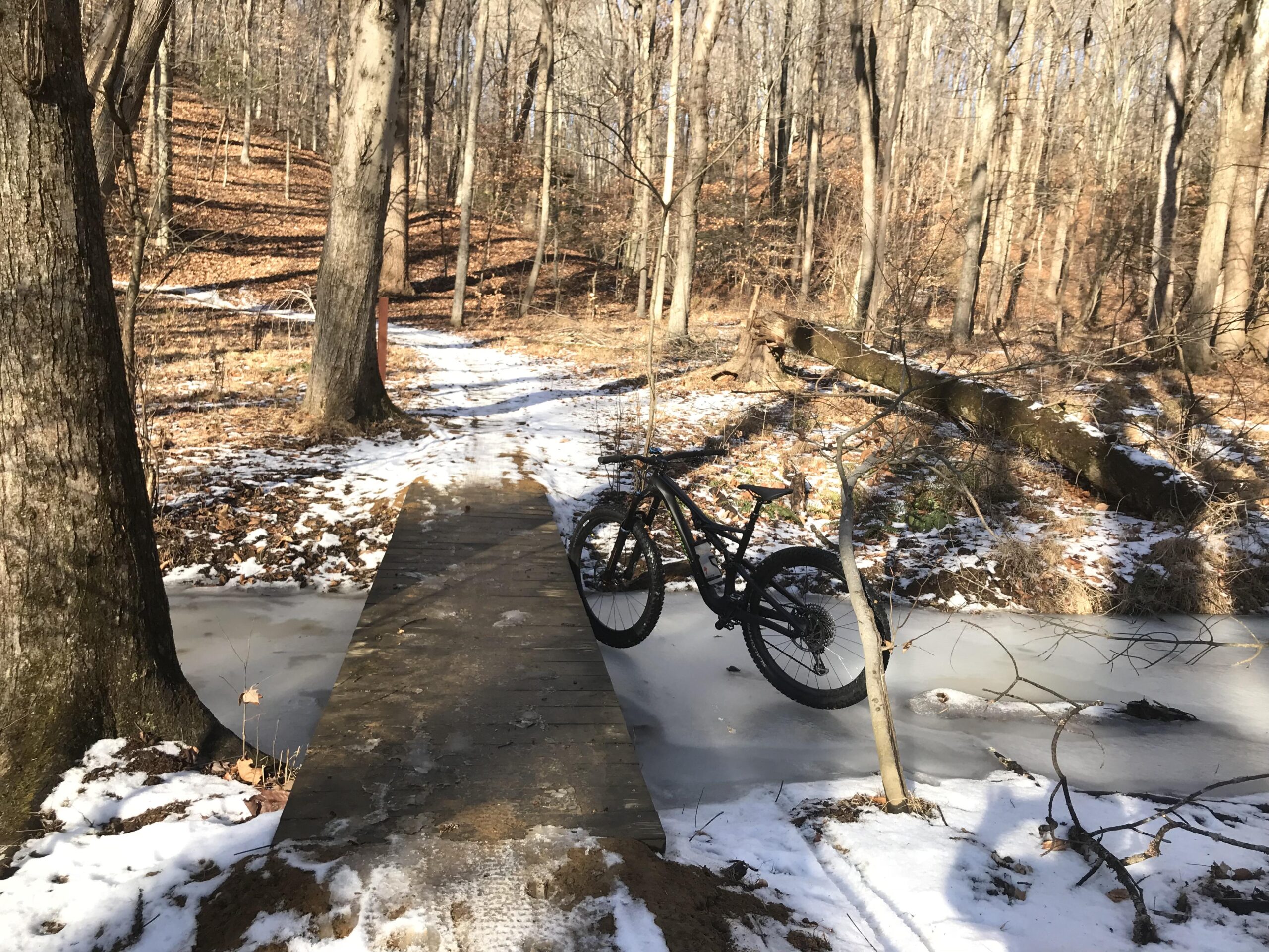 Specialized Stumpjumper S Works FSR: A mountain bike resting on a narrow wooden bridge over a frozen stream, surrounded by a wooded area with bare trees and patches of snow on the ground. A dirt path winds through the forest, leading into the distance.