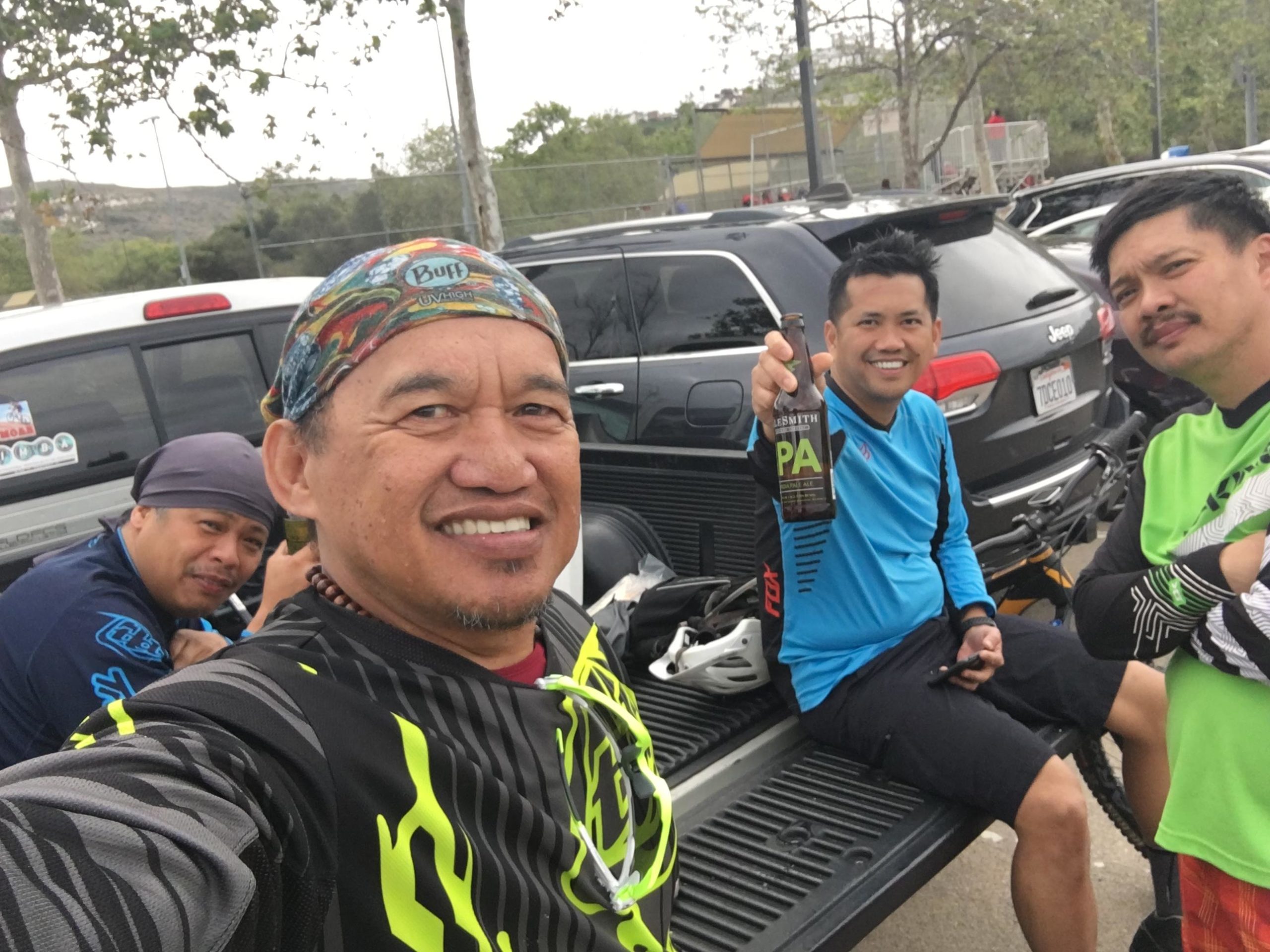 A group of four friends enjoying a day outdoors, sitting on the tailgate of a truck in a parking area. They are dressed in vibrant athletic clothing, with two individuals wearing helmets. One person is holding a bottle of beer, while others are smiling at the camera. Trees and a sports facility are visible in the background, suggesting a recreational setting. Los Penasquitos Canyon Preserve mountain bike trail.