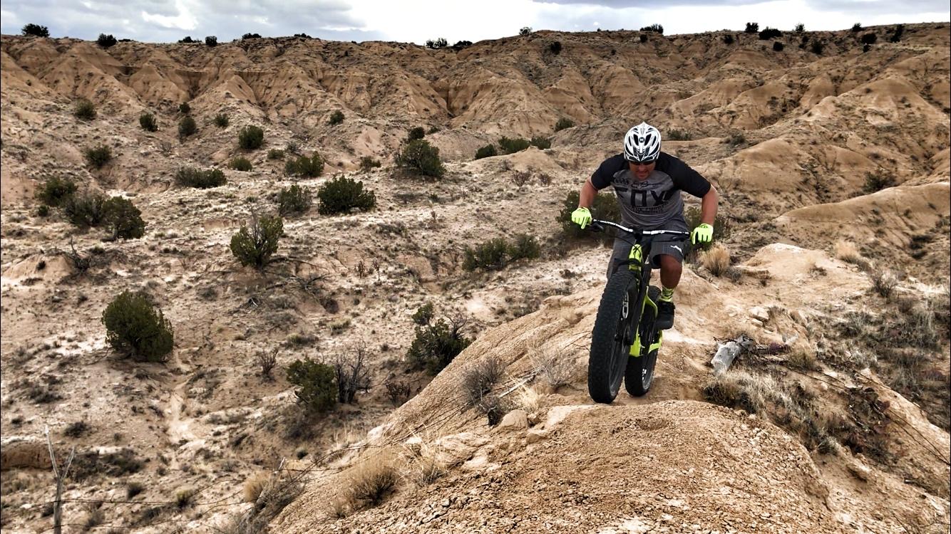 A mountain biker navigating a rocky hillside, performing a stunt on a rugged terrain. The landscape features dry, sandy hills and sparse vegetation under a cloudy sky. The biker is wearing a helmet and gear, and the bike has large tires designed for off-road riding. Mariposa Fat Bike Trails mountain bike trail.