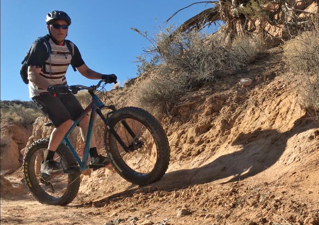 A cyclist riding a fat bike on a rugged dirt trail. The biker is wearing a helmet and cycling gear, navigating a steep incline surrounded by sparse vegetation and rocky terrain under a clear blue sky. Mariposa Fat Bike Trails mountain bike trail.
