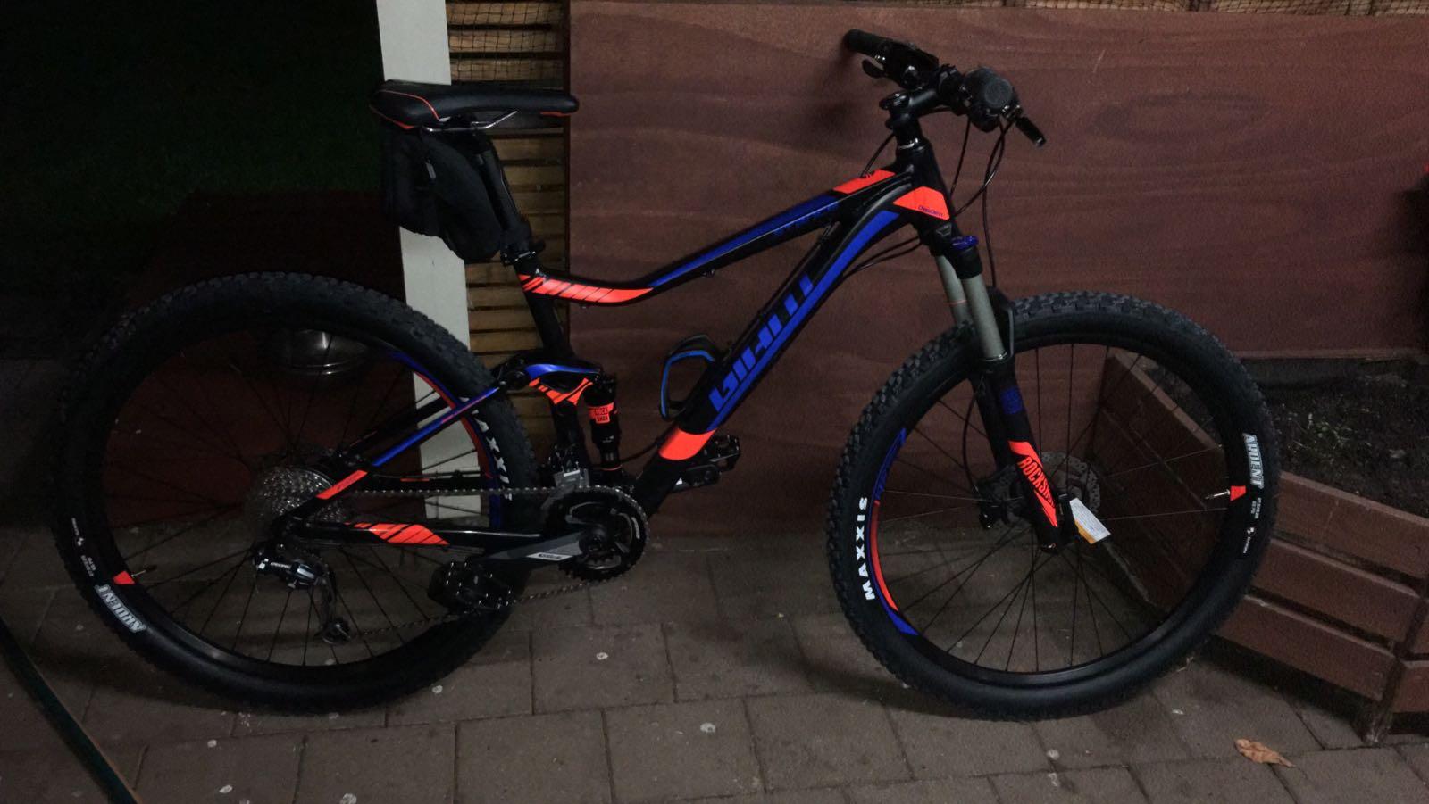 Giant Giant Trance Advanced 27.5 2: A mountain bike with a black and orange frame is leaning against a wooden wall. The bike features thick tires, a visible gear system, and a small black storage bag attached to the seat. The background shows a dimly lit outdoor area with wooden panels and some foliage.