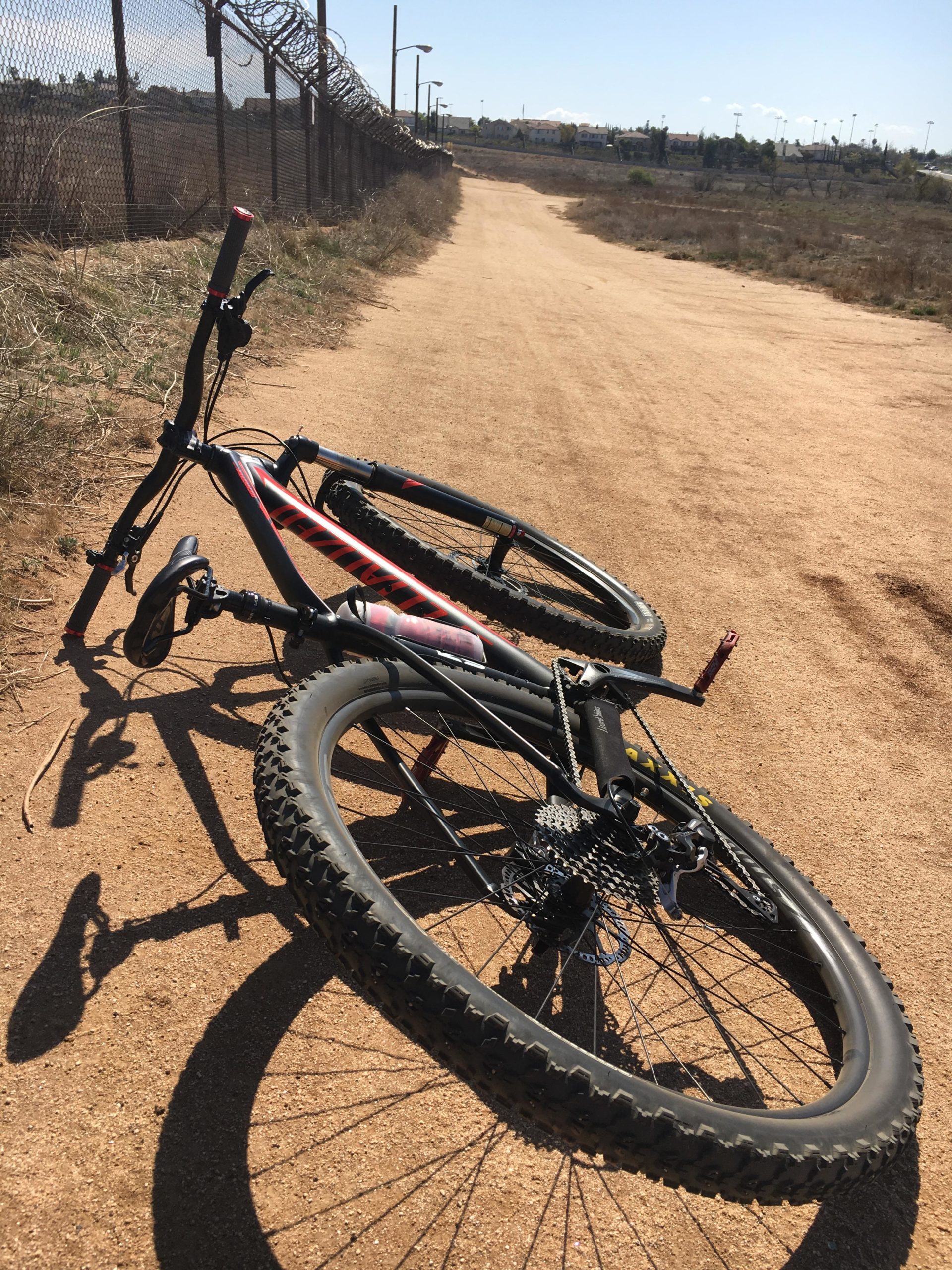 A mountain bike lying on its side on a sandy path, with a barbed wire fence in the background. The scene is under a clear blue sky, and there is sparse vegetation along the trail. Sycamore Canyon Park mountain bike trail.