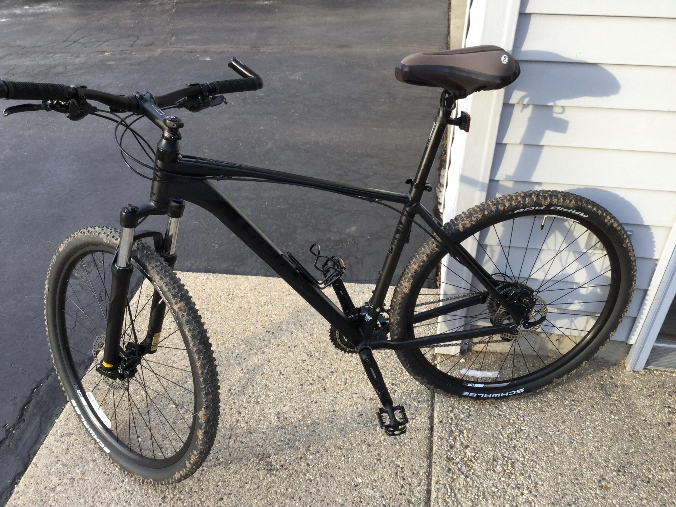 Giant Talon 29er: A black mountain bike leaning against a wall, showcasing its front suspension, knobby tires with dirt residue, and black handlebars. The bike is positioned on a concrete surface with a portion of a building visible in the background.