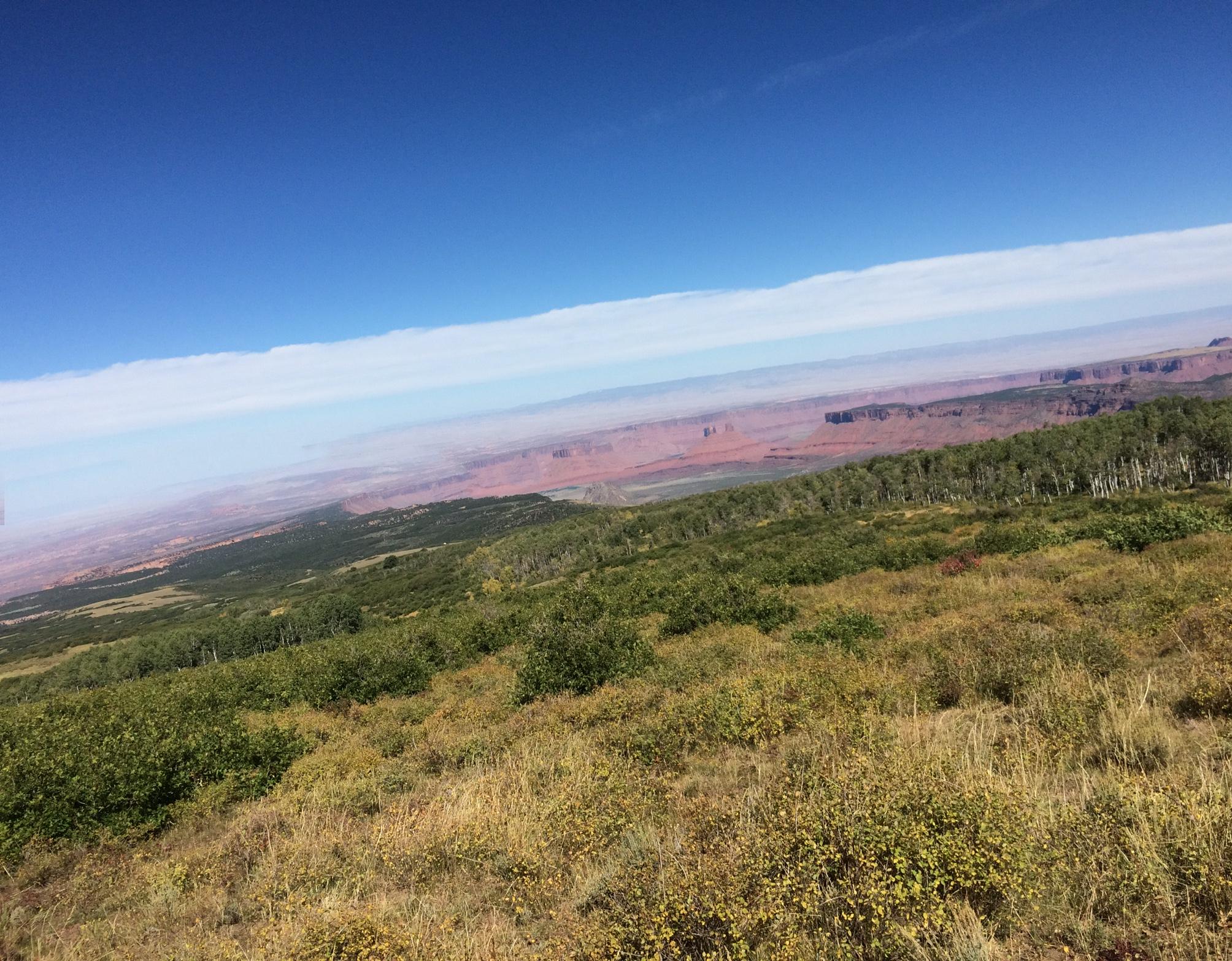 A panoramic view of a mountainous landscape featuring green vegetation in the foreground, with rolling hills leading to distant red rock formations under a clear blue sky. The Whole Enchilada mountain bike trail.