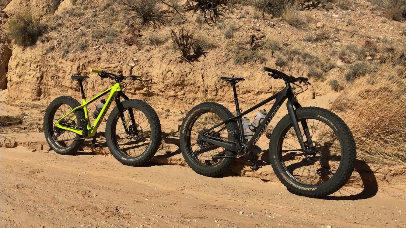Two fat tire bikes are parked on a sandy trail, with a rocky hillside in the background. The bike on the left is bright green, while the one on the right is black with a design. Both bikes have large, rugged tires, indicating they're suitable for off-road cycling. The environment appears dry and natural, featuring sparse vegetation and rocky terrain. Mariposa Fat Bike Trails mountain bike trail.