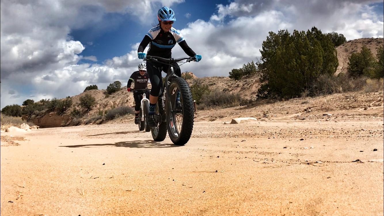A female cyclist riding a fat bike on a sandy trail, with a male cyclist visible in the background. The scene takes place in a rugged outdoor environment under a partly cloudy sky, surrounded by sparse vegetation and rocky terrain. Mariposa Fat Bike Trails mountain bike trail.