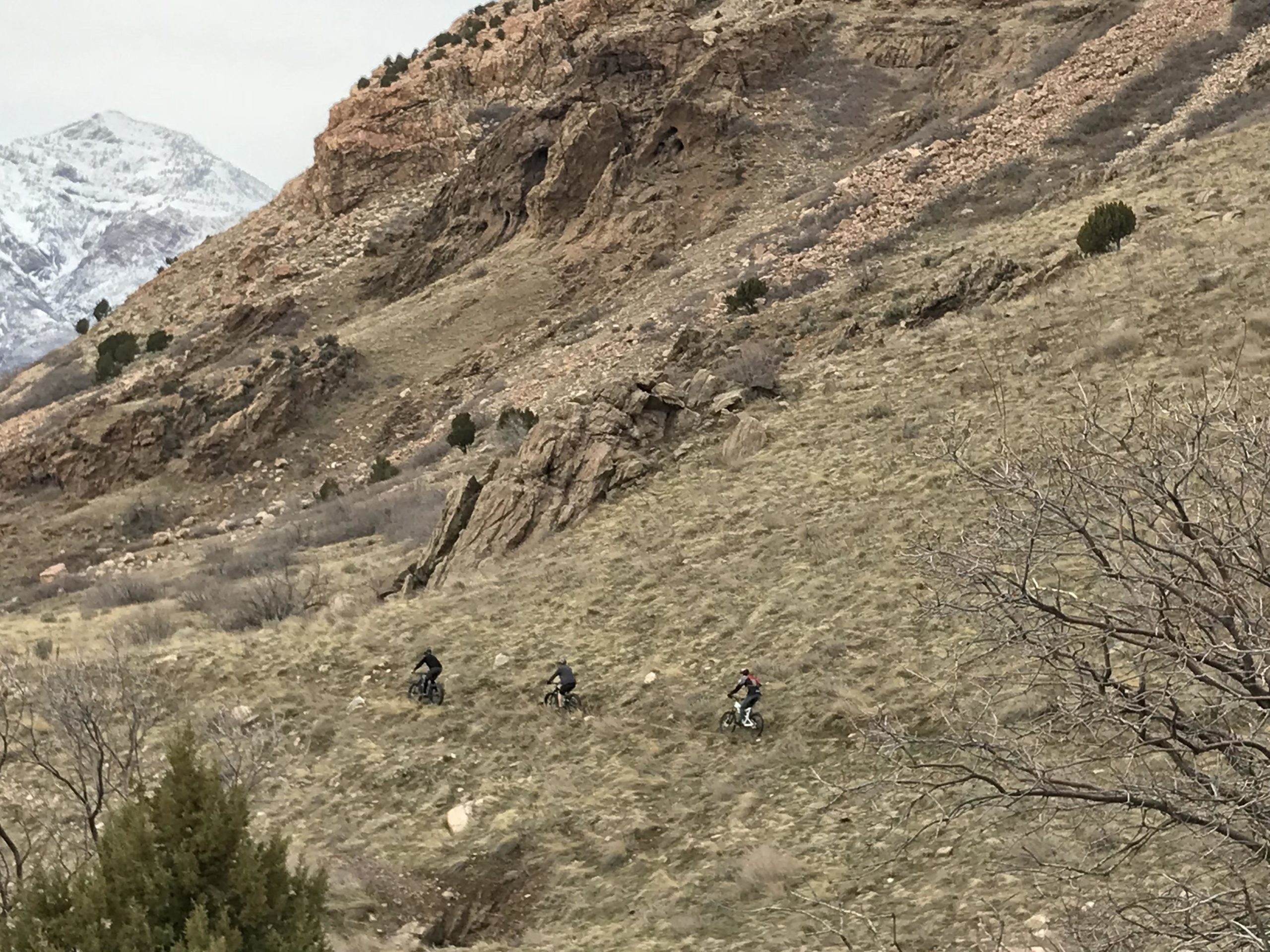 Three mountain bikers navigating a grassy hillside with rocky terrain and snow-capped mountains in the background. The scene is set in a natural landscape with sparse vegetation and rugged features, under a cloudy sky. Bonneville Shoreline Ogden North Of 12th mountain bike trail.