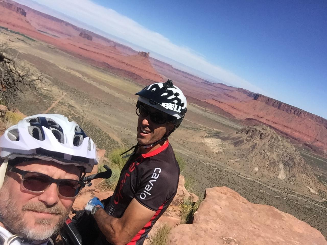 A selfie of two mountain bikers wearing helmets, set against a scenic backdrop of red rock formations and a vast desert landscape under a clear blue sky. One biker appears to be adjusting their bike, while the other smiles directly at the camera. The Whole Enchilada mountain bike trail.