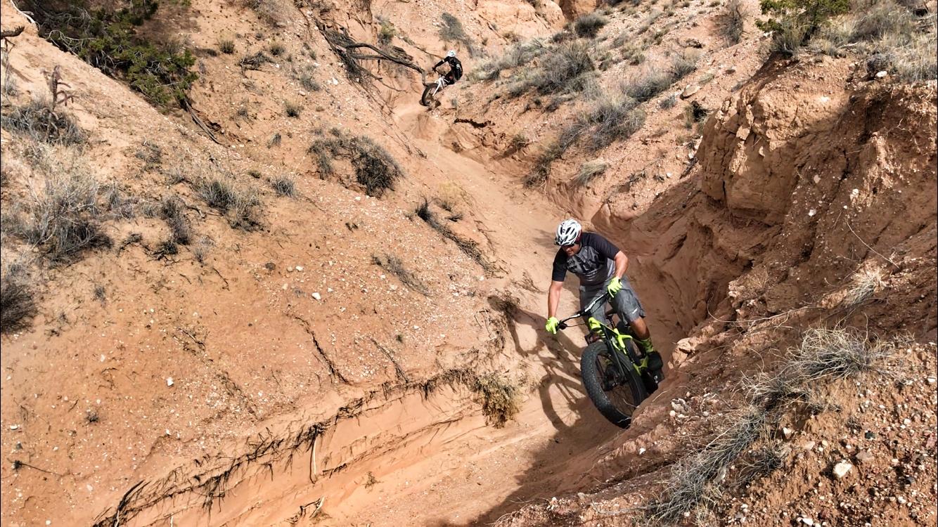 Two mountain bikers navigating a narrow dirt trail in a rocky desert landscape. One biker is descending a steep, sandy path, while the other is visible in the background. The terrain features reddish soil and sparse vegetation, typical of arid environments. Mariposa Fat Bike Trails mountain bike trail.