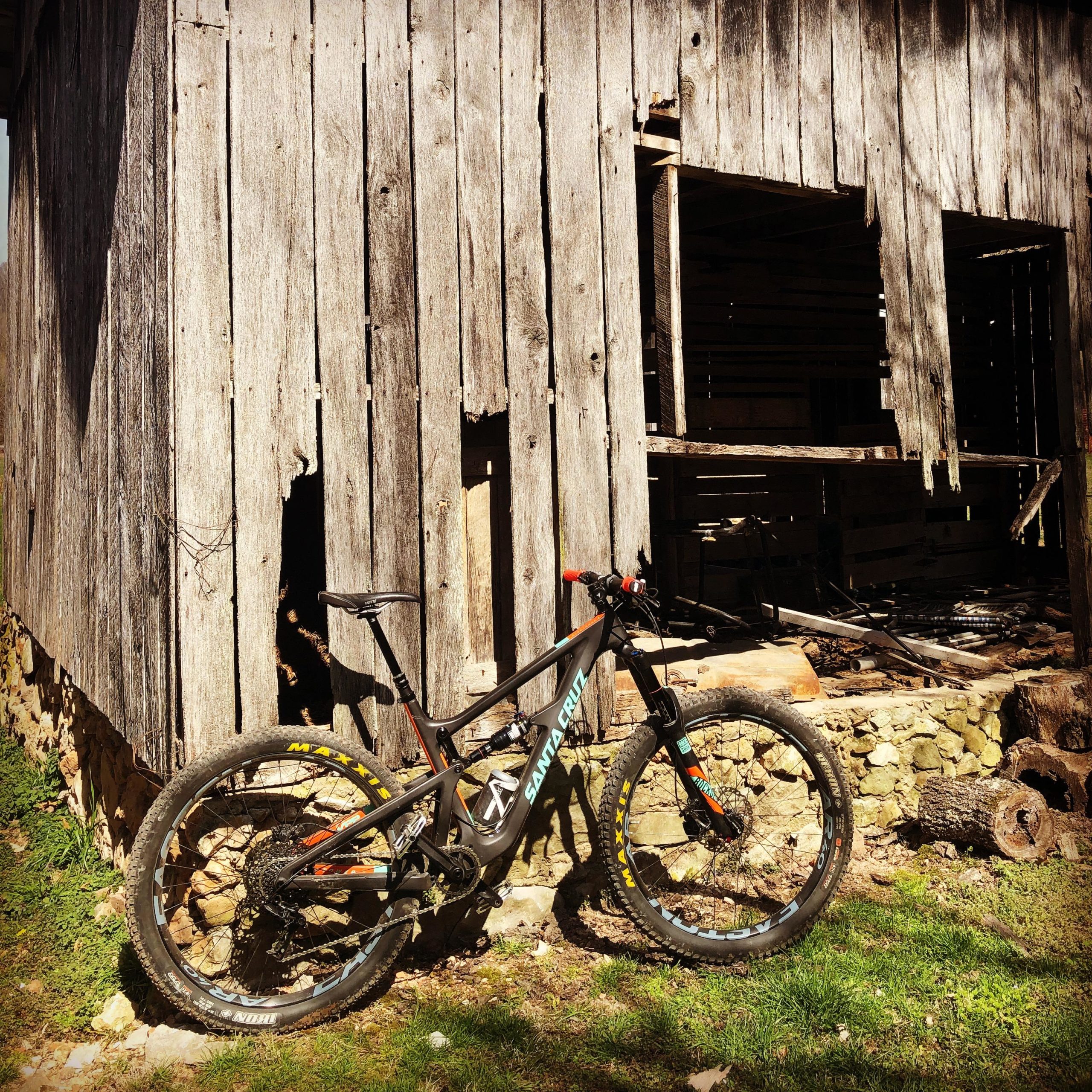 A mountain bike leaning against a rustic, weathered wooden barn with an open window and a partially collapsed wall. The surrounding area features grass and an assortment of logs and stones. The scene captures a blend of nature and abandoned structure, evoking a sense of adventure and exploration. Coler Mountain Bike Preserve mountain bike trail.