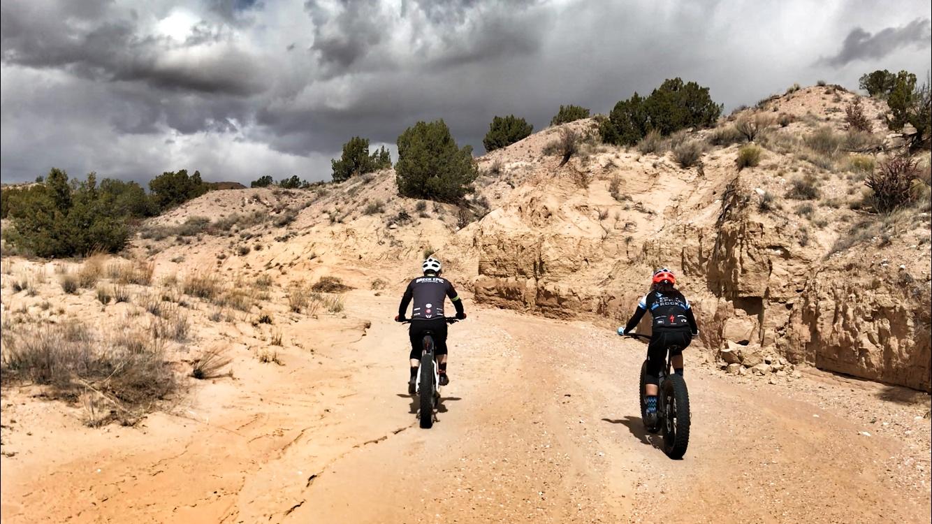 Two cyclists riding fat tire bikes along a sandy dirt trail in a rugged, arid landscape, with sparse vegetation and rocky hills under a cloudy sky. Mariposa Fat Bike Trails mountain bike trail.