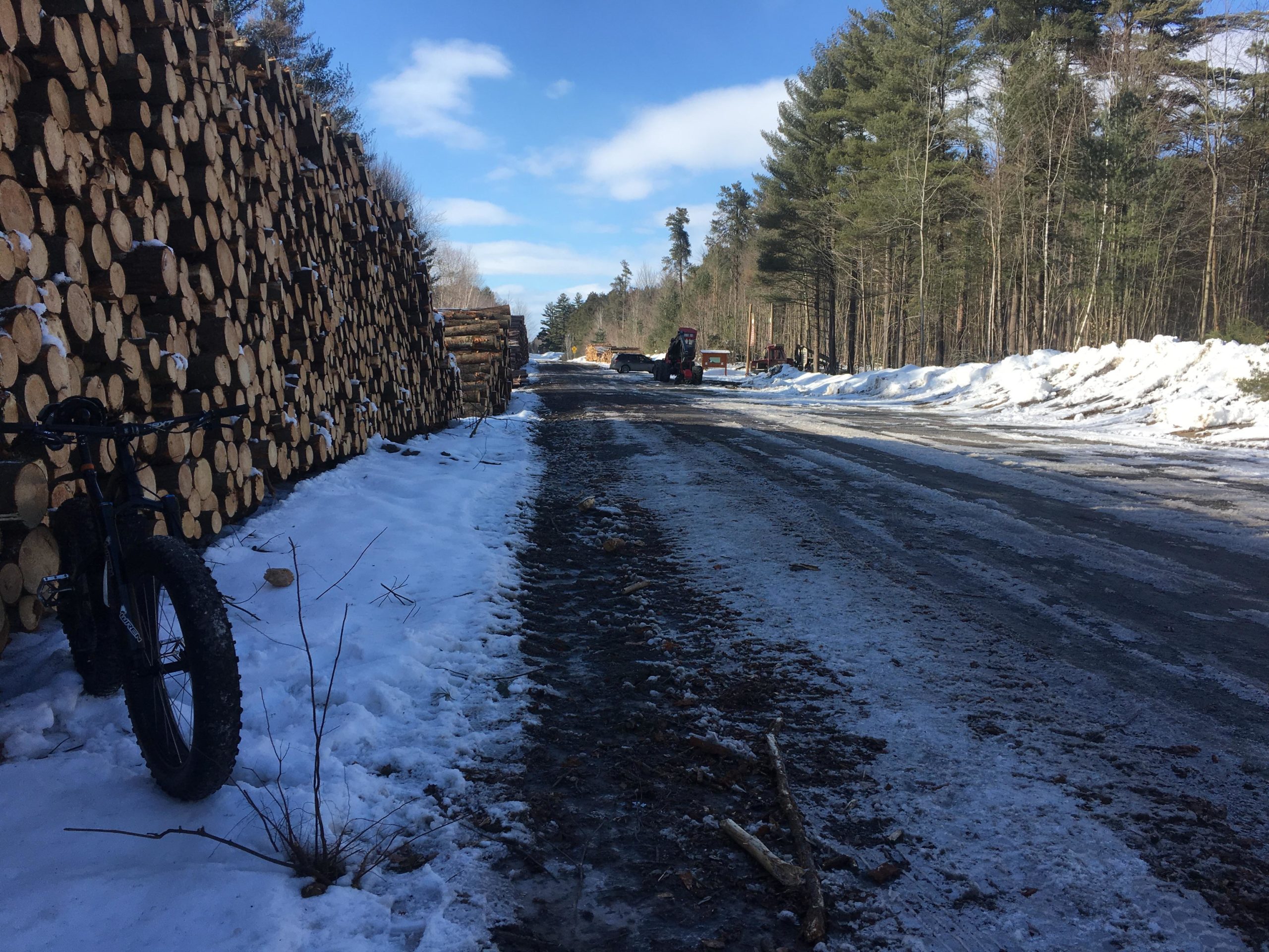 A snowy forest road lined with stacked logs on one side. A fat-tire bicycle is parked on the snow-covered edge, while a tractor is visible in the distance, working near the edge of the tree line. The scene is set against a backdrop of clear blue skies and scattered clouds. Larose Forest mountain bike trail.