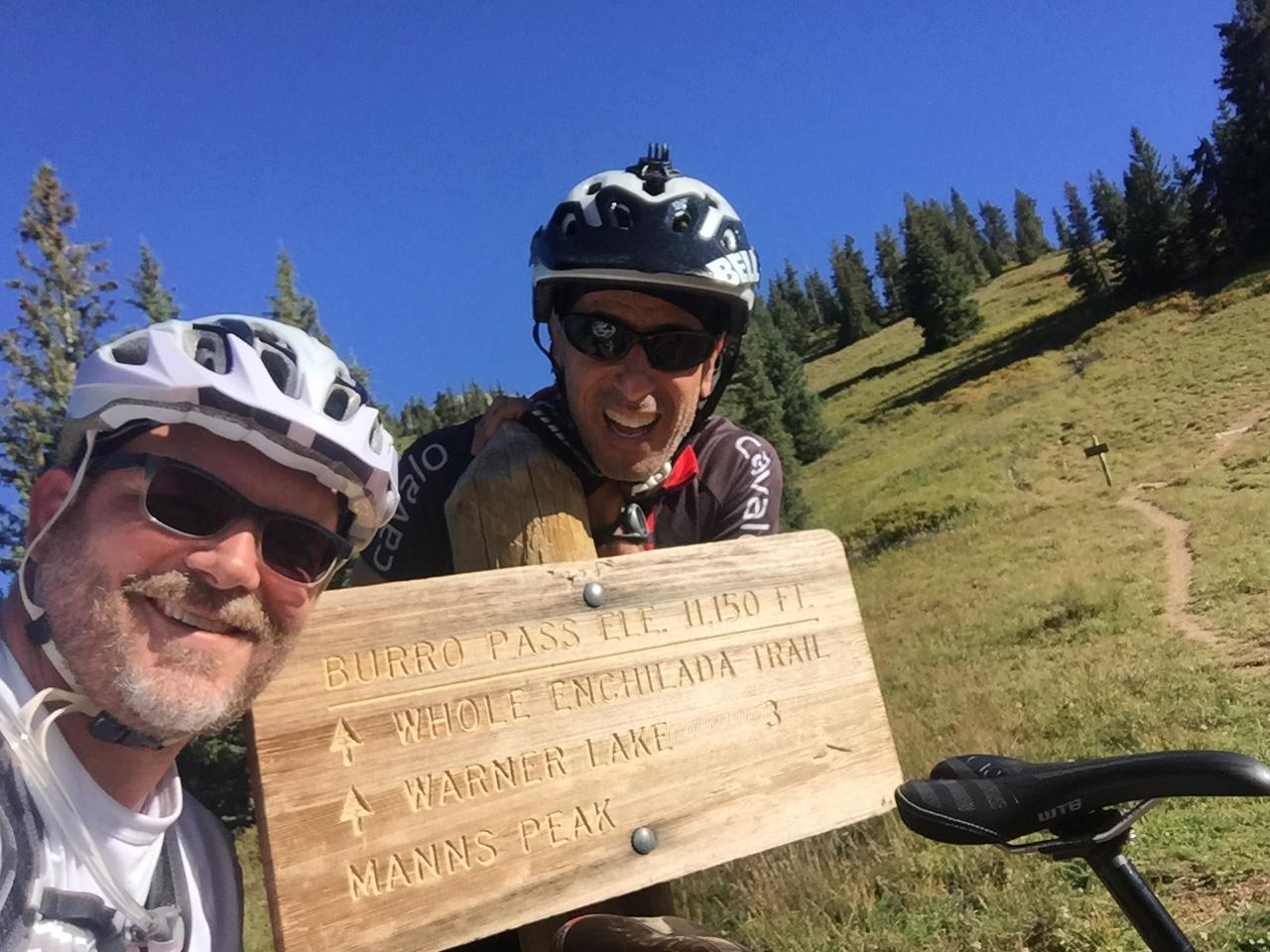 Two cyclists are smiling in front of a wooden trail sign that reads "Burro Pass Elev. 11,150 ft." The sign points to various hiking trails, including "Whole Enchilada Trail," "Warner Lake," and "Mann's Peak." In the background, there are lush green hills and trees under a clear blue sky. The Whole Enchilada mountain bike trail.
