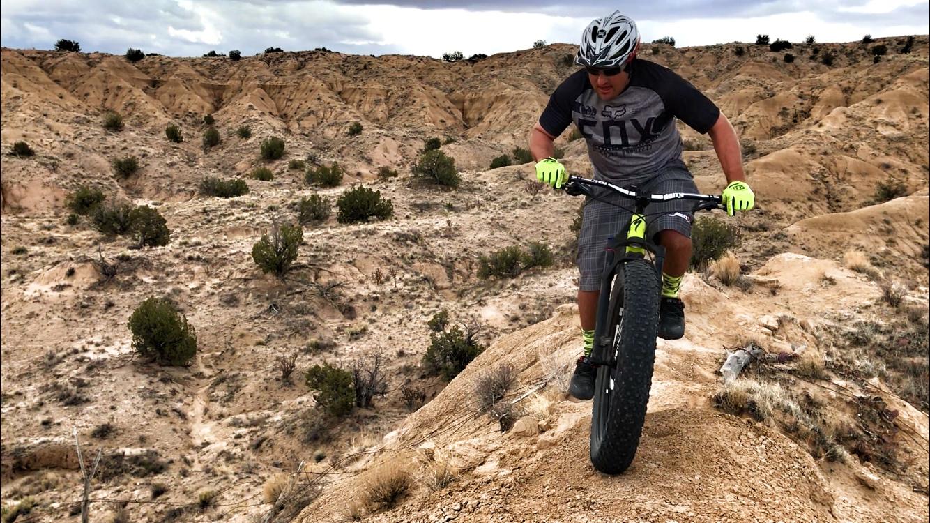 A mountain biker navigating a rocky terrain, wearing a helmet and brightly colored gloves, with rugged hills and sparse vegetation in the background. The cyclist is focused on maneuvering the bike down a slope. Mariposa Fat Bike Trails mountain bike trail.