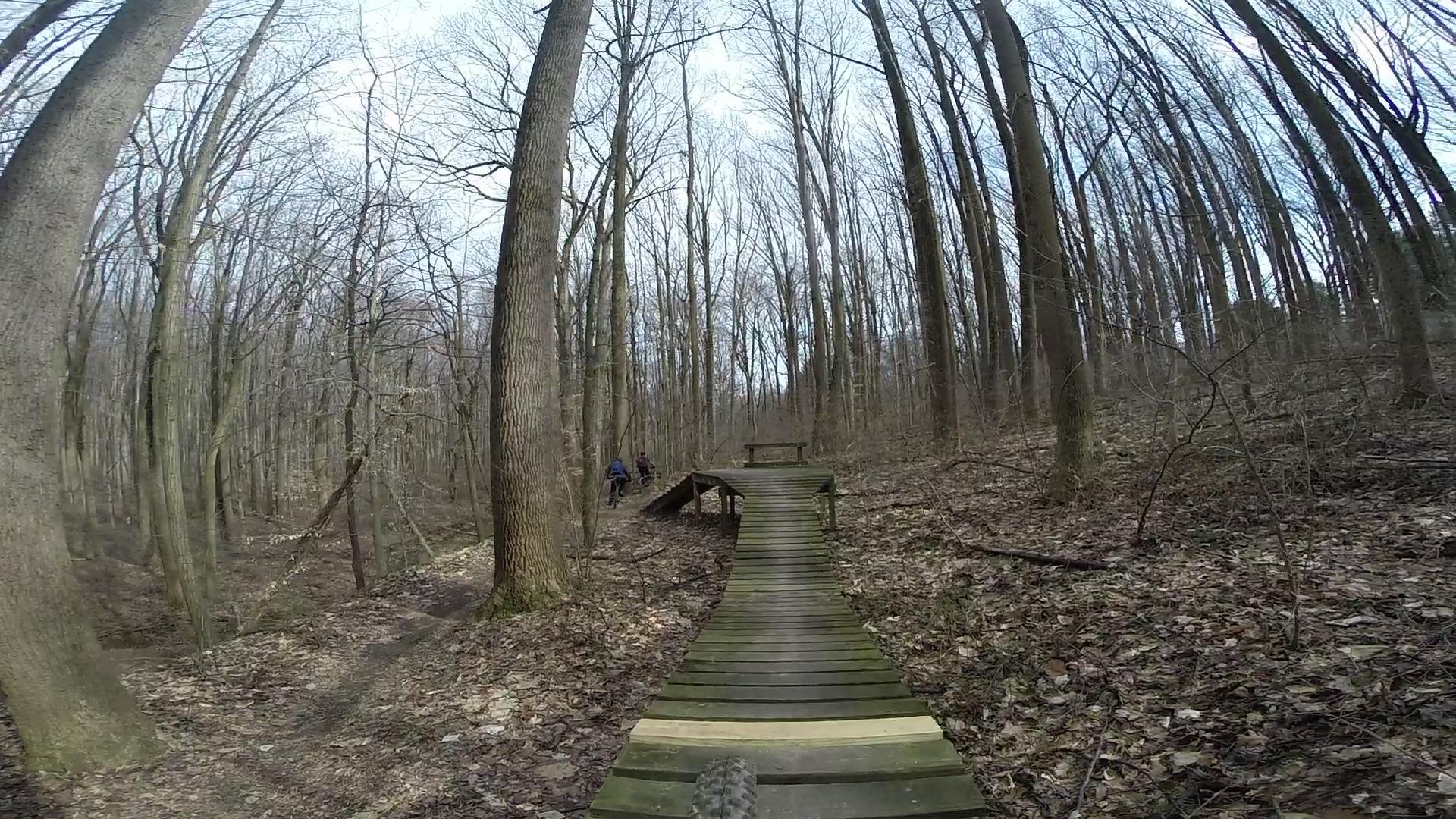 A wooden path leads through a forest with bare trees in early spring, while two individuals can be seen biking in the background. The ground is covered with fallen leaves, creating a natural trail. The sky above is partly cloudy. White Clay Creek mountain bike trail.