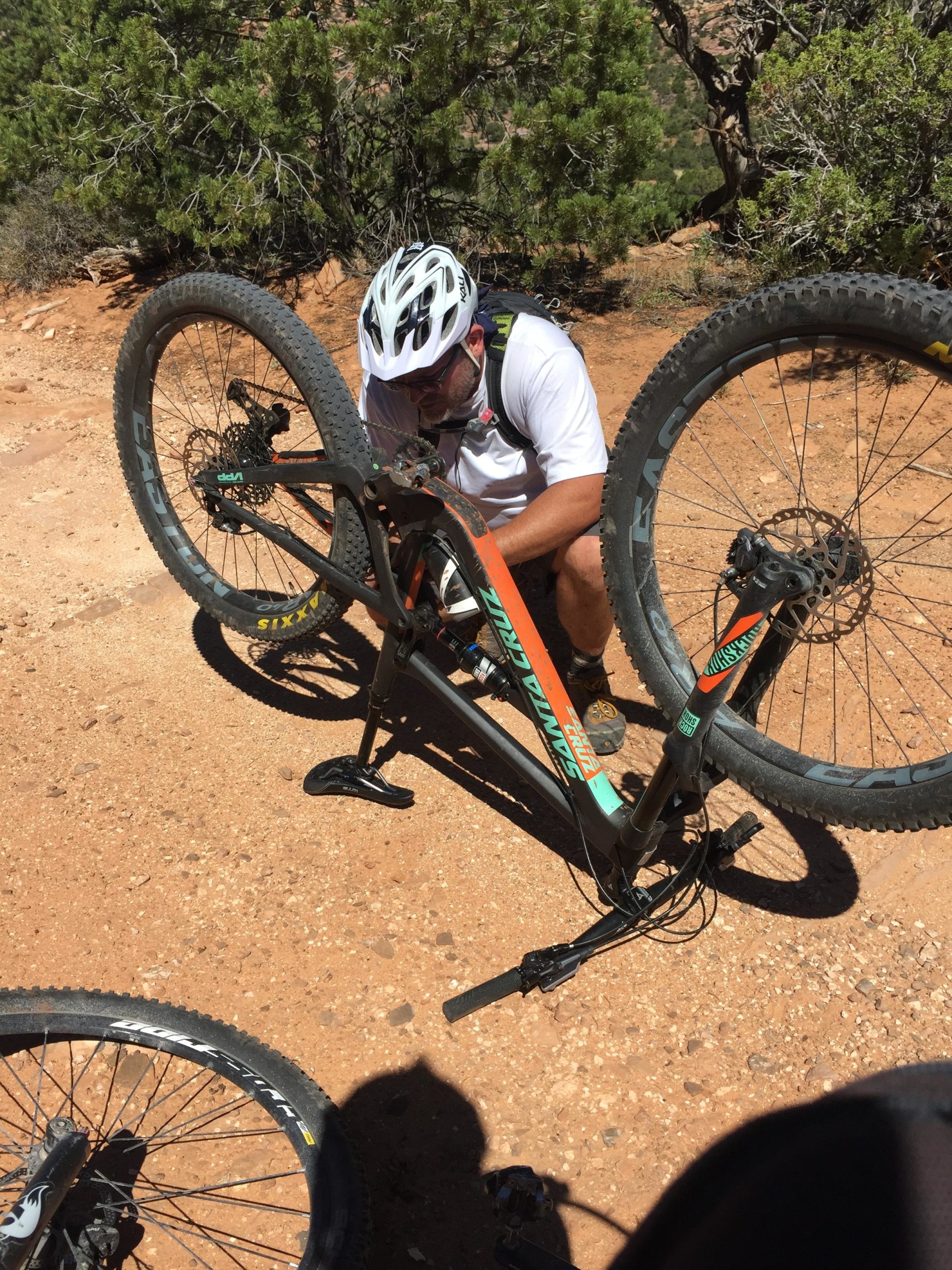 A cyclist in a helmet is kneeling beside a mountain bike with its rear wheel lifted off the ground, inspecting it on a dirt path surrounded by shrubs and rocky terrain. The scene captures a moment of bike maintenance during an outdoor ride. The Whole Enchilada mountain bike trail.