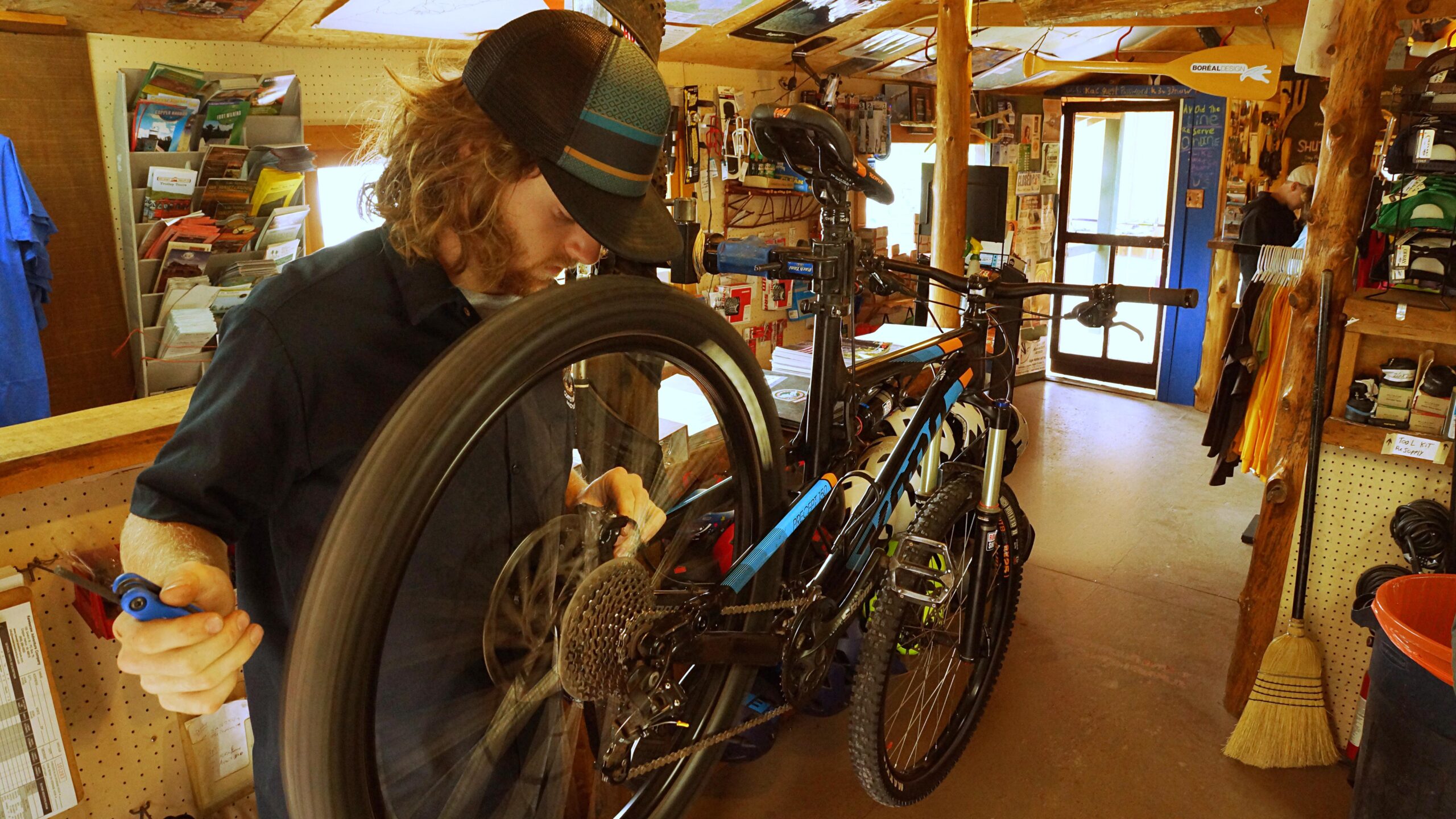 A young man wearing a cap is focused on repairing a mountain bike's rear wheel in a bike shop. The background features various biking equipment and promotional materials, with natural wood accents in the shop's decor.