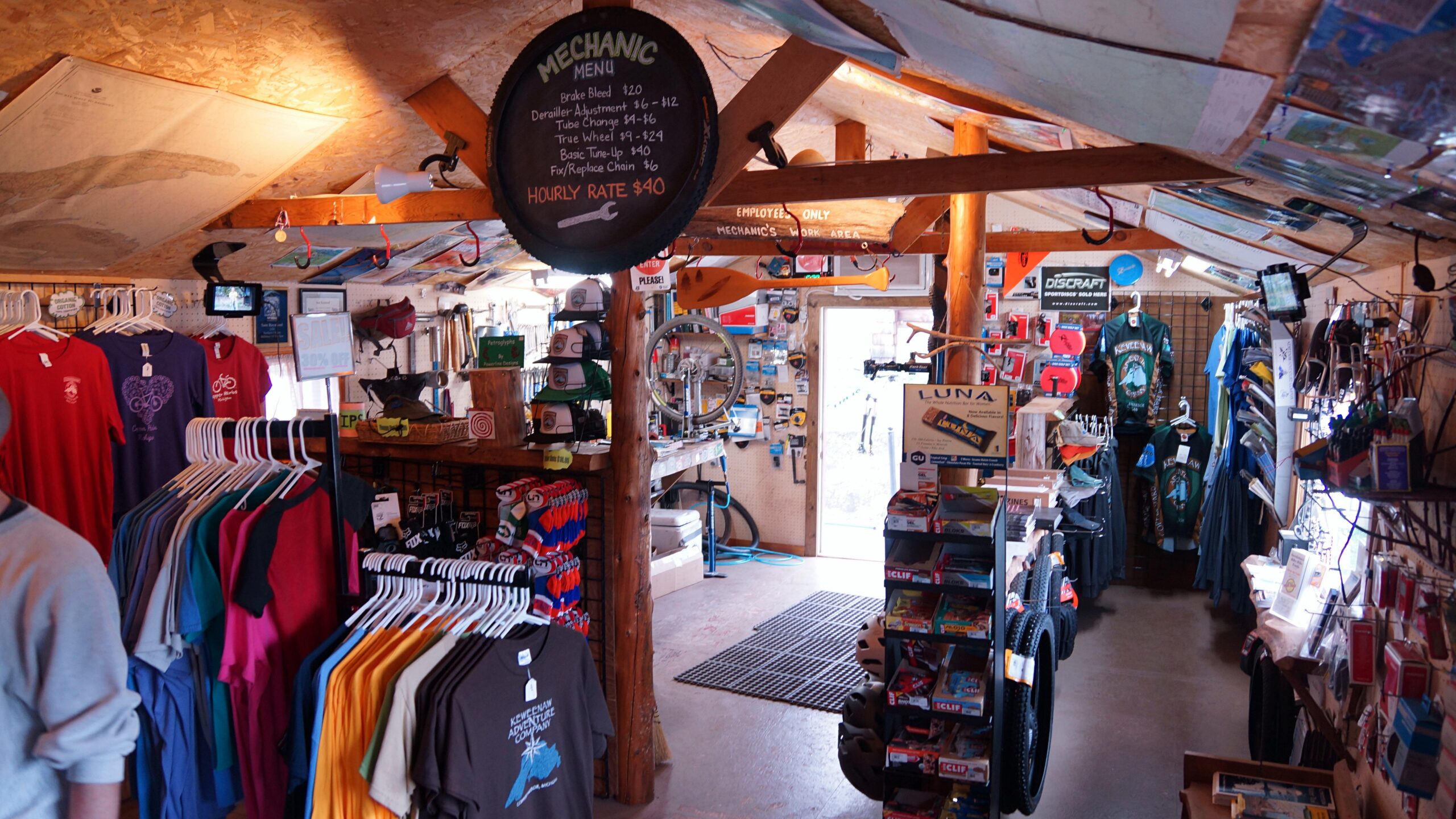 Interior of a small shop featuring a variety of merchandise, including colorful t-shirts displayed on racks, outdoor gear, and bike repair tools. A chalkboard displays a mechanic menu with pricing for services. The shop is warmly lit with wooden beams, and there is a door leading outside visible in the background.